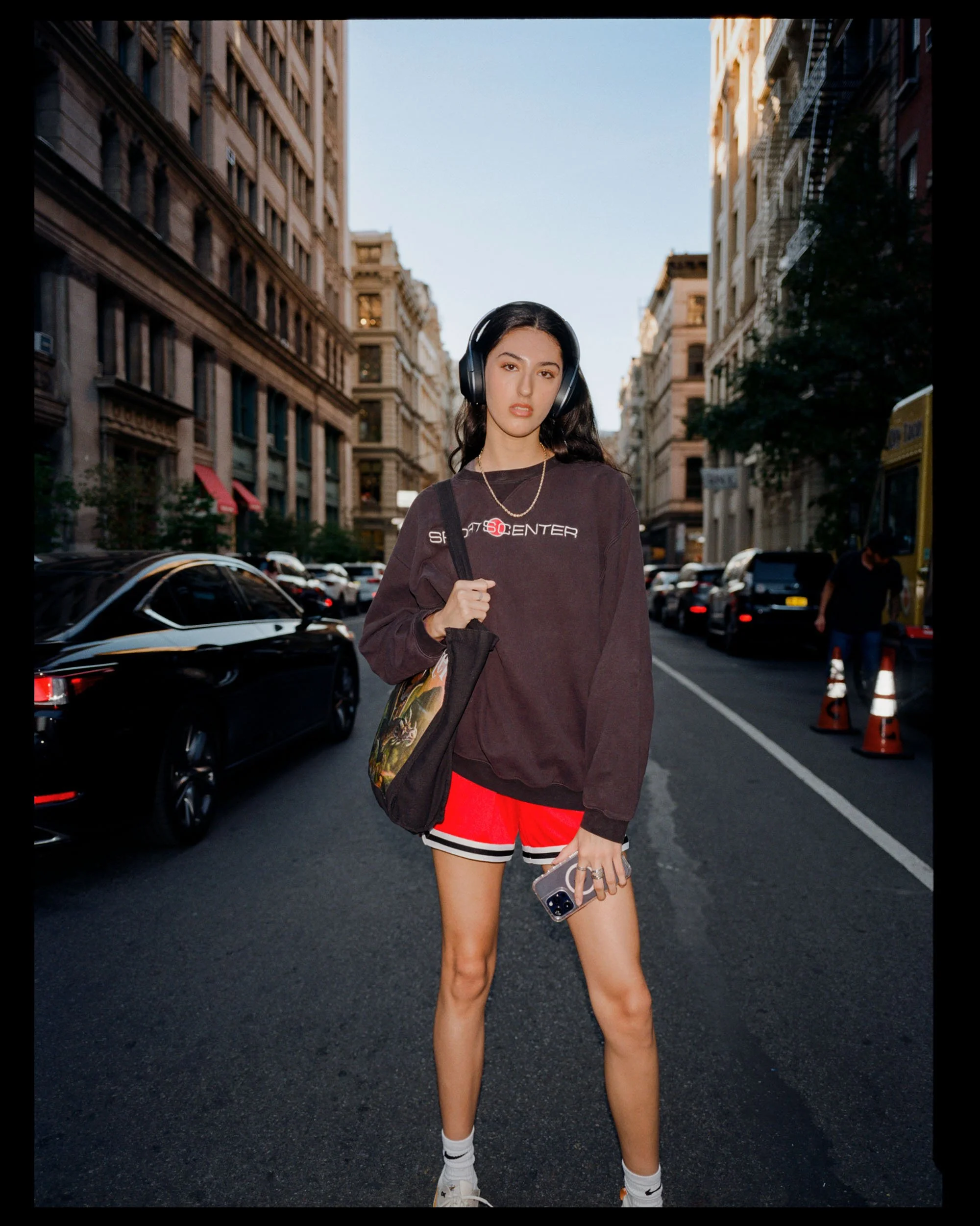 A young woman wearing headphones, a dark oversized sweatshirt, red and white shorts, and sneakers, standing on a city street with tall buildings, parked cars, and traffic cones visible in the background