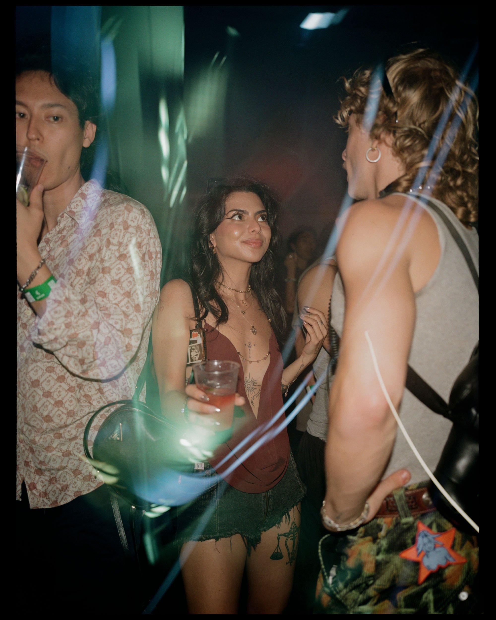 People socializing at a nightclub or party, with focus on a woman holding a drink and looking between two other women, all dressed casually, in a dimly lit environment.