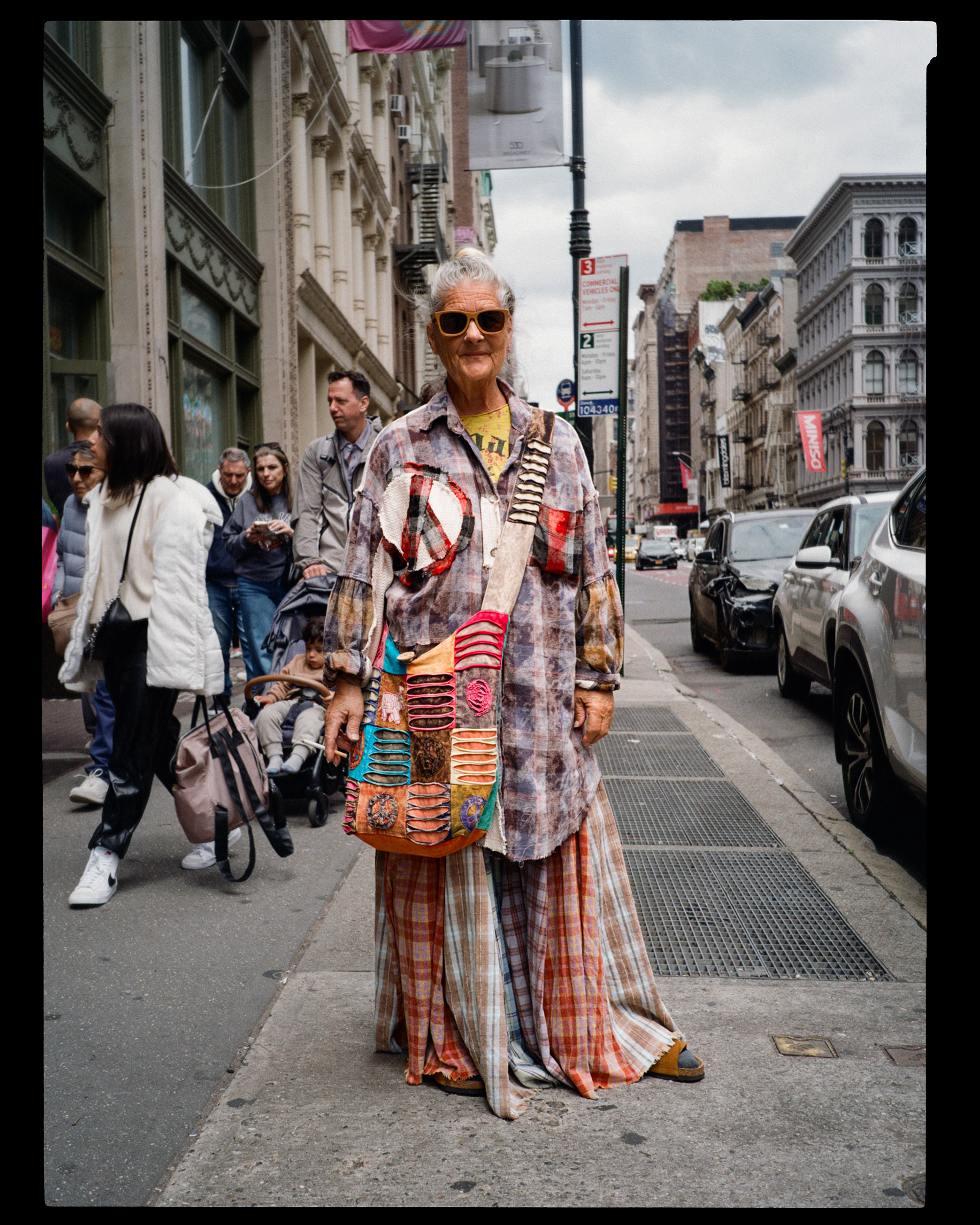 An elderly woman in colorful, patchwork clothing and sunglasses standing on a city sidewalk, with a crowd of people behind her and cars parked along the street.