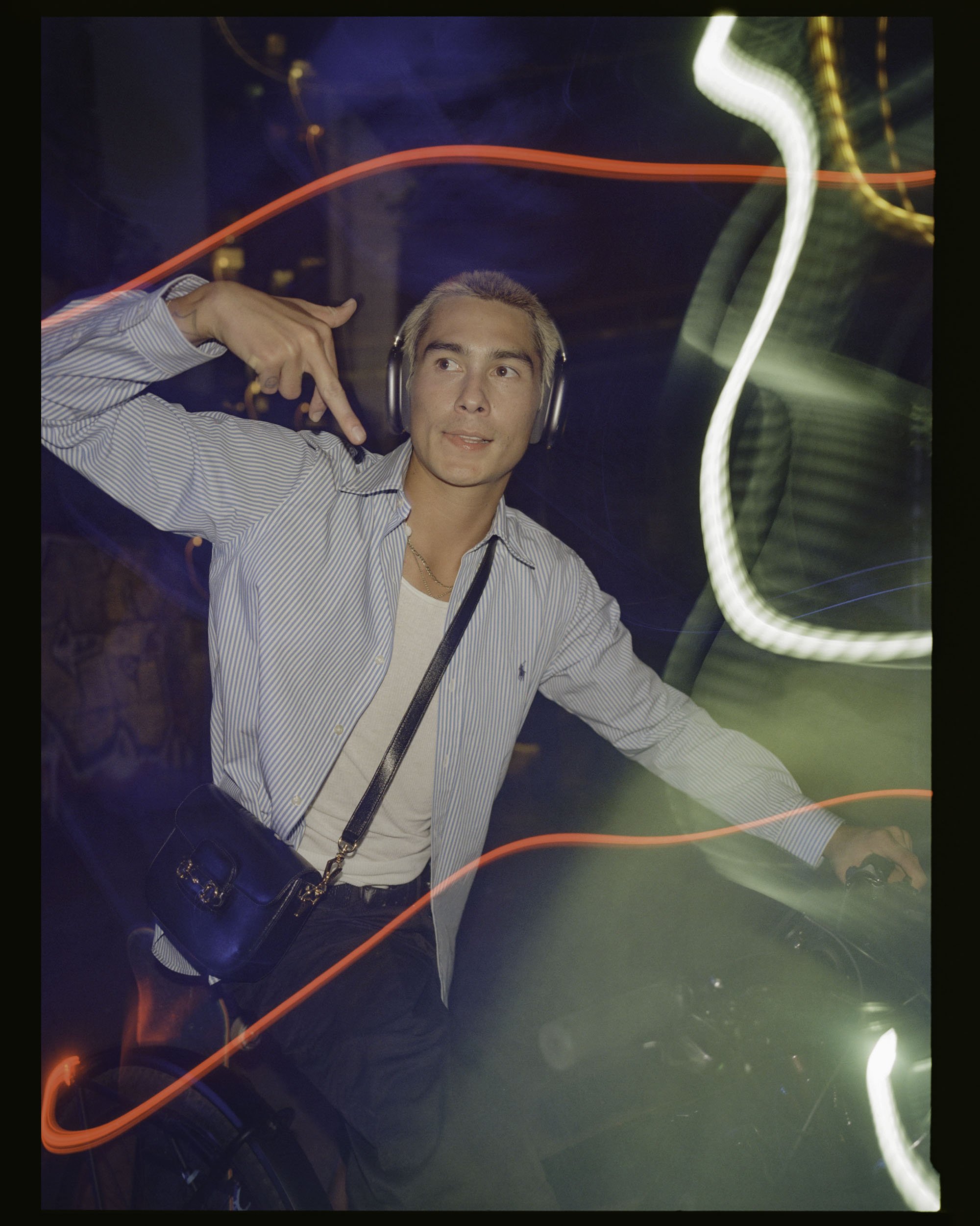 A young man with blonde hair wearing headphones, a striped shirt, and a white undershirt, making a peace sign gesture with his right hand while looking to the side. The background features colorful light trails and abstract lighting effects.