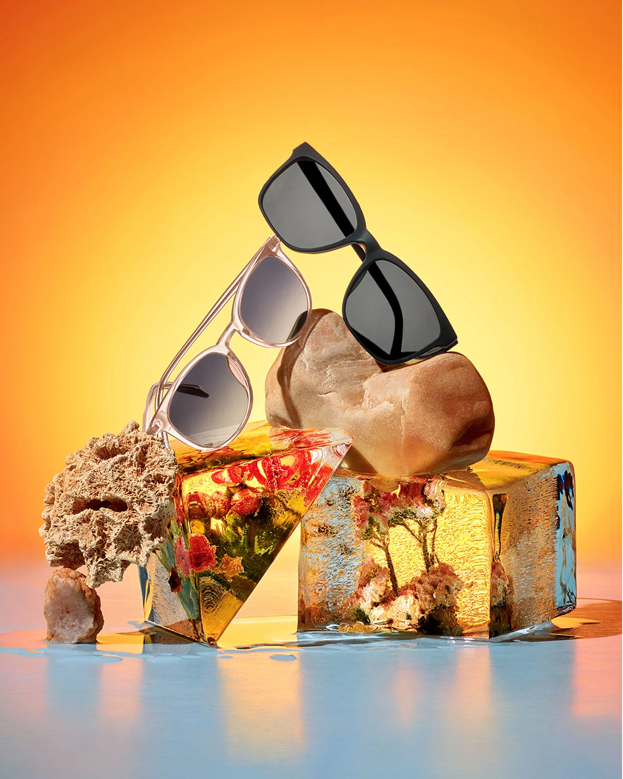 A collection of sunglasses positioned on a rock and colorful geometric blocks with an orange gradient background and water reflection.