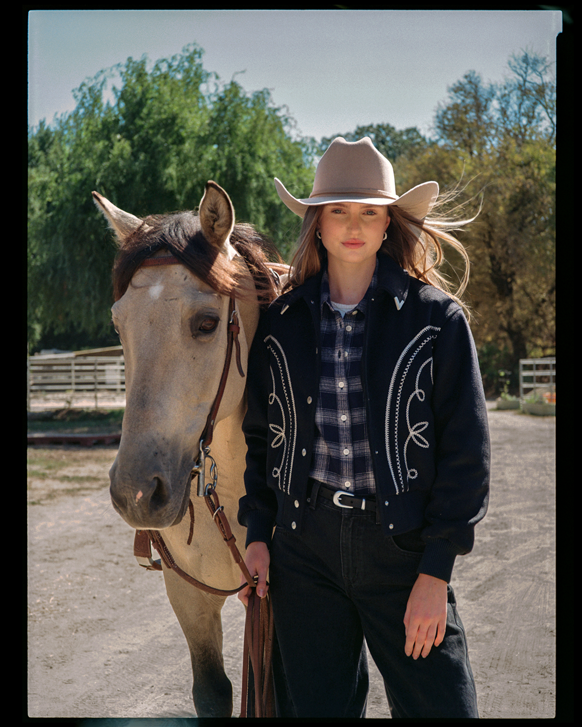 A woman wearing a large beige cowboy hat and a dark jacket with embroidery, standing outside on a sunny day with a horse beside her. The horse is light-colored with a white blaze on its face and a brown mane. There are trees and a wooden fence in the