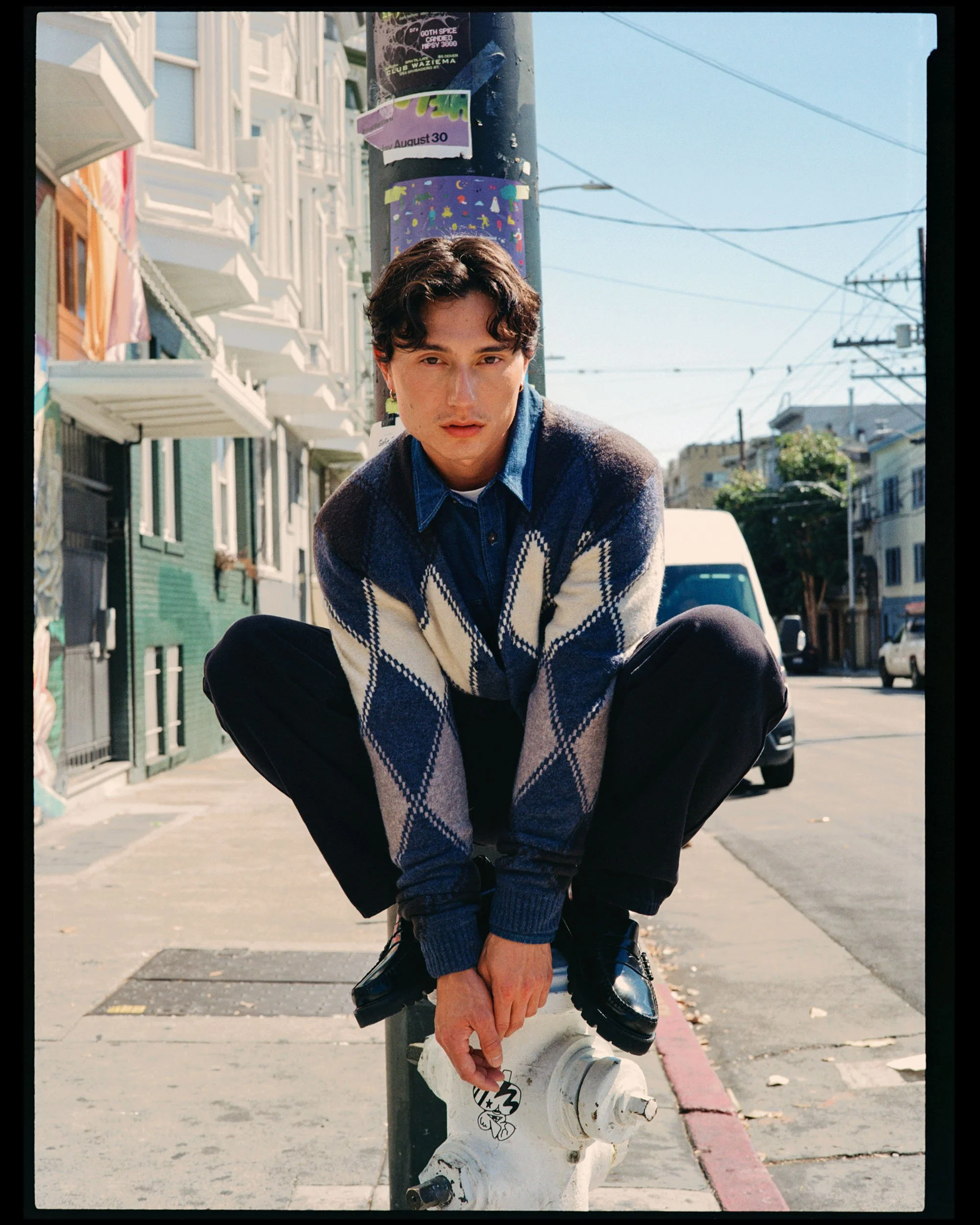 A young man with curly hair wearing a blue checkered sweater, sitting on a fire hydrant on a city street, looking at the camera, with buildings, power lines, and parked cars in the background.