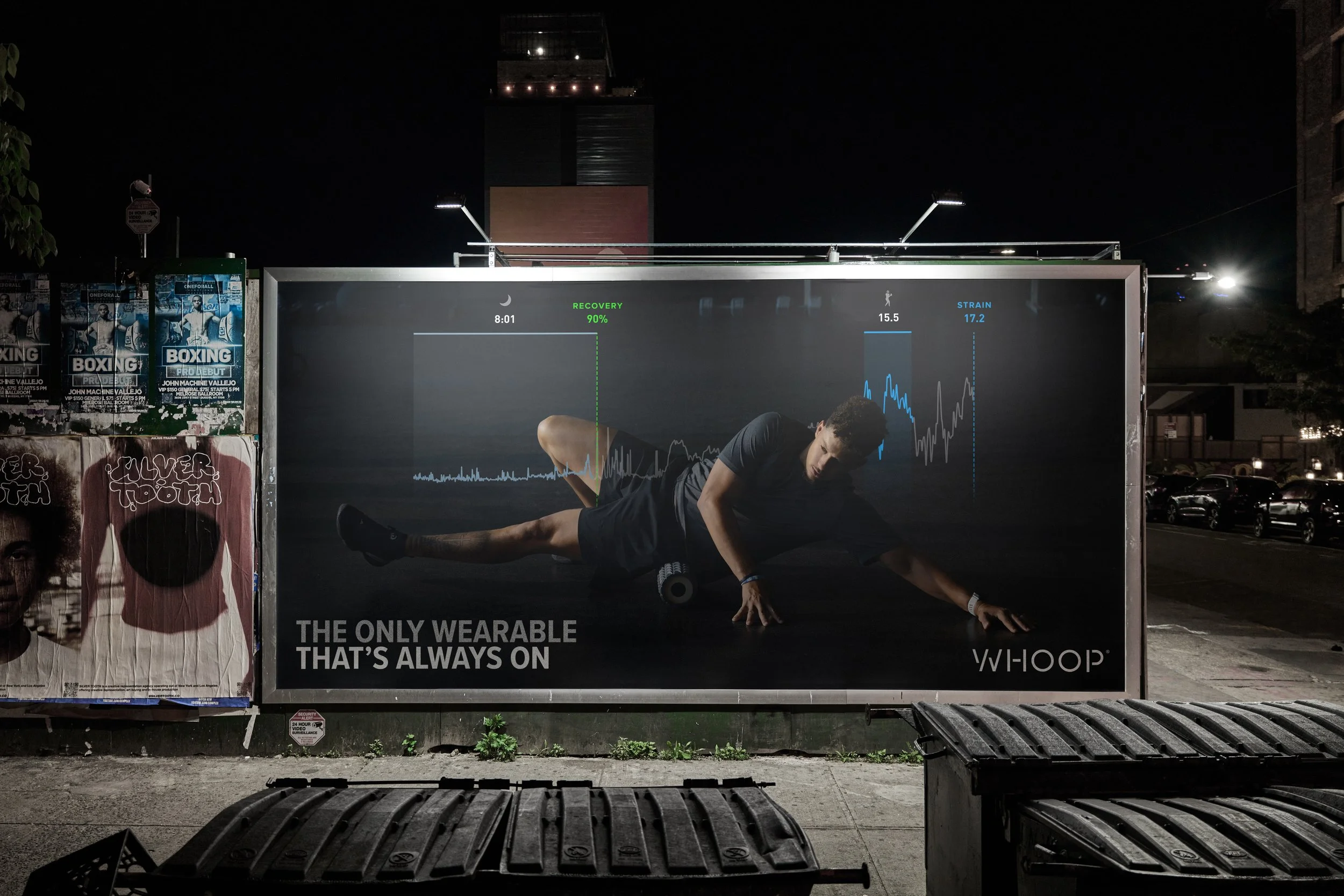 A billboard at night displays a man doing a stretch exercise on a fitness tracker called WHOOP, with graphs showing recovery, strain, and sleep data. The billboard reads 'The only wearable that's always on'.