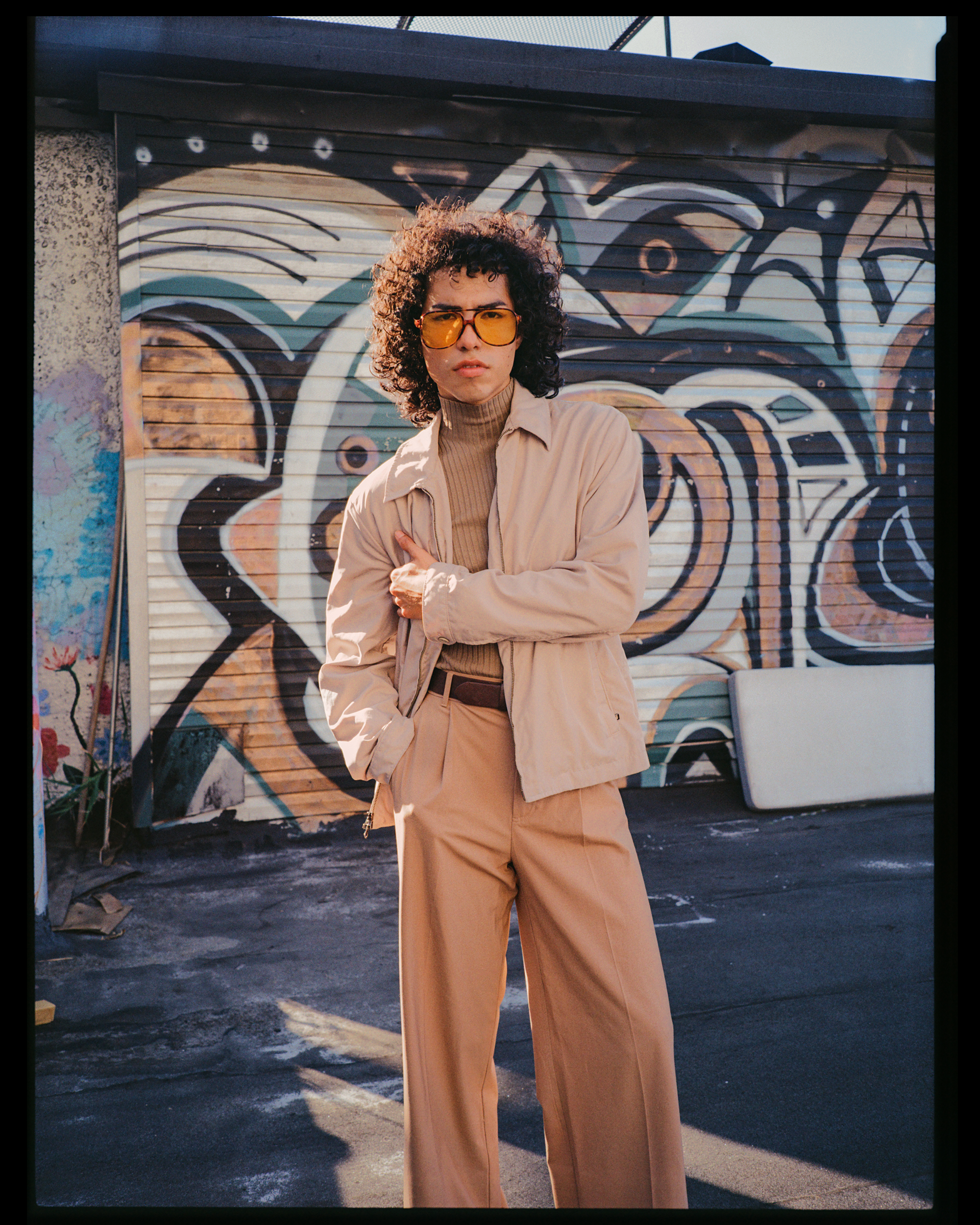 A woman with curly hair wearing yellow-tinted sunglasses, a tan jacket, and beige pants, standing with her arms crossed in front of a colorful graffiti mural on a metal garage door.