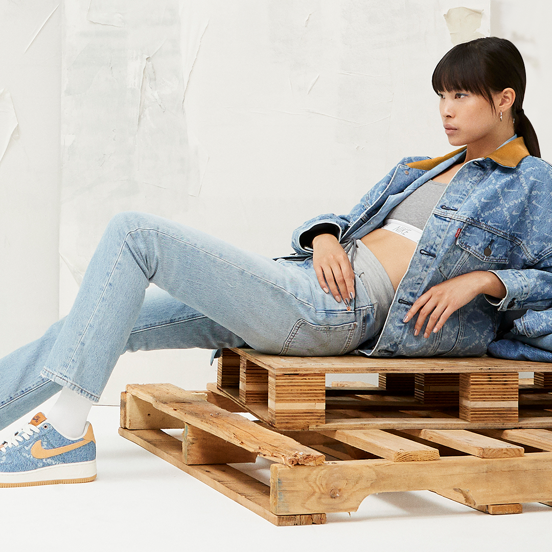 Young woman reclining on a wooden pallet with casual denim clothing, Nike sneakers, and dark hair with bangs, inside a minimalistic room with a white wall background.