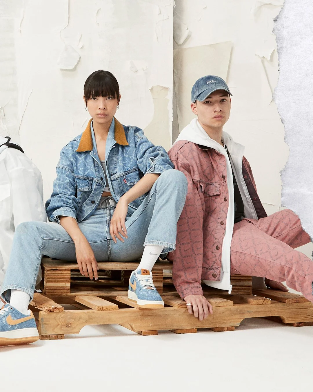 Two young people sitting on a wooden pallet in a studio with a torn wall background. The woman on the left is wearing a denim jacket with a brown collar, light blue jeans, and Nike sneakers. The man on the right is wearing a pink patterned jacket, a white hoodie, pink patterned pants, and a blue cap.