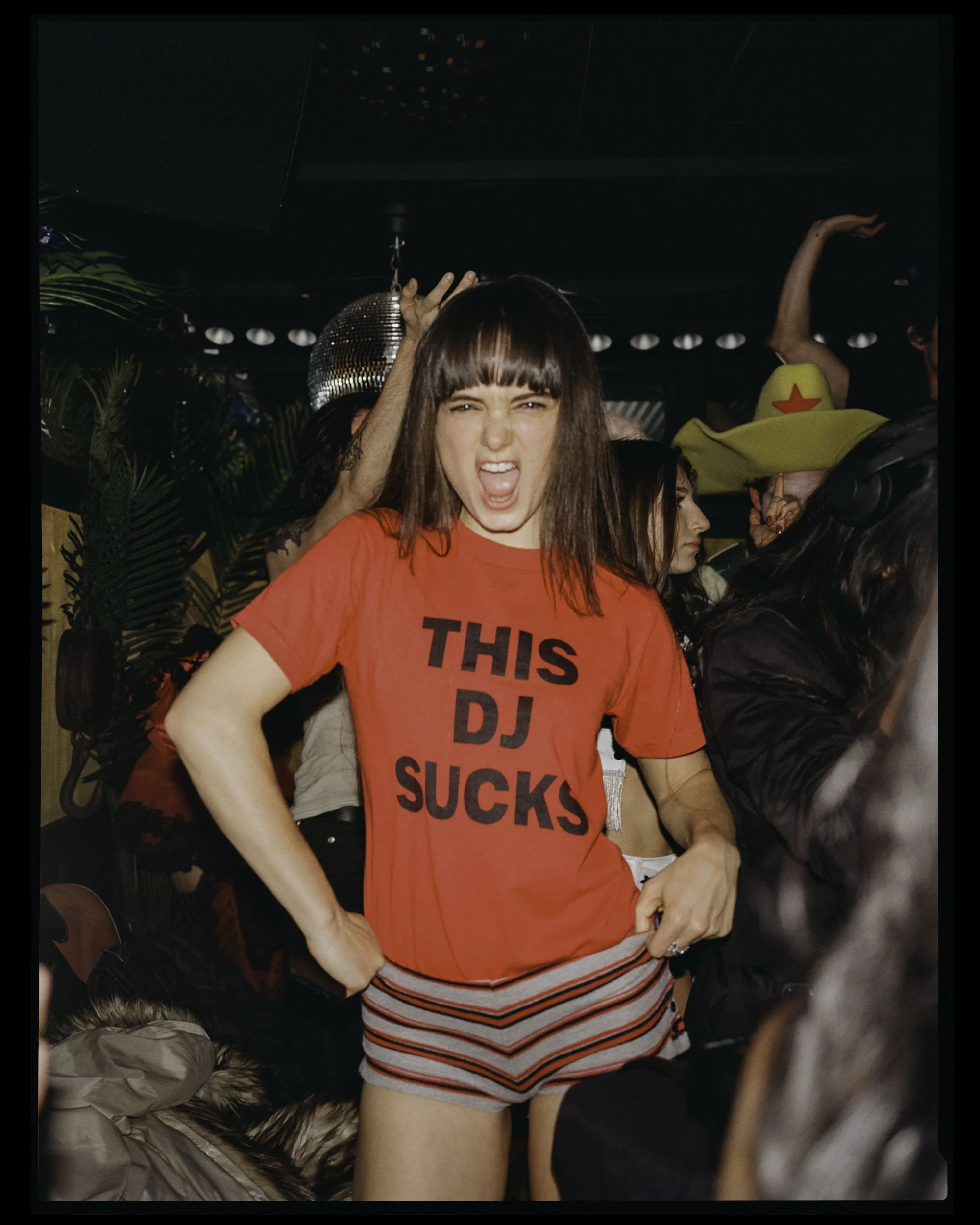 A young woman with dark hair and bangs, wearing a red T-shirt with black text and striped shorts, standing in a crowded party or club scene. She has a playful, rebellious expression, sticking out her tongue and making a peace sign with her right hand