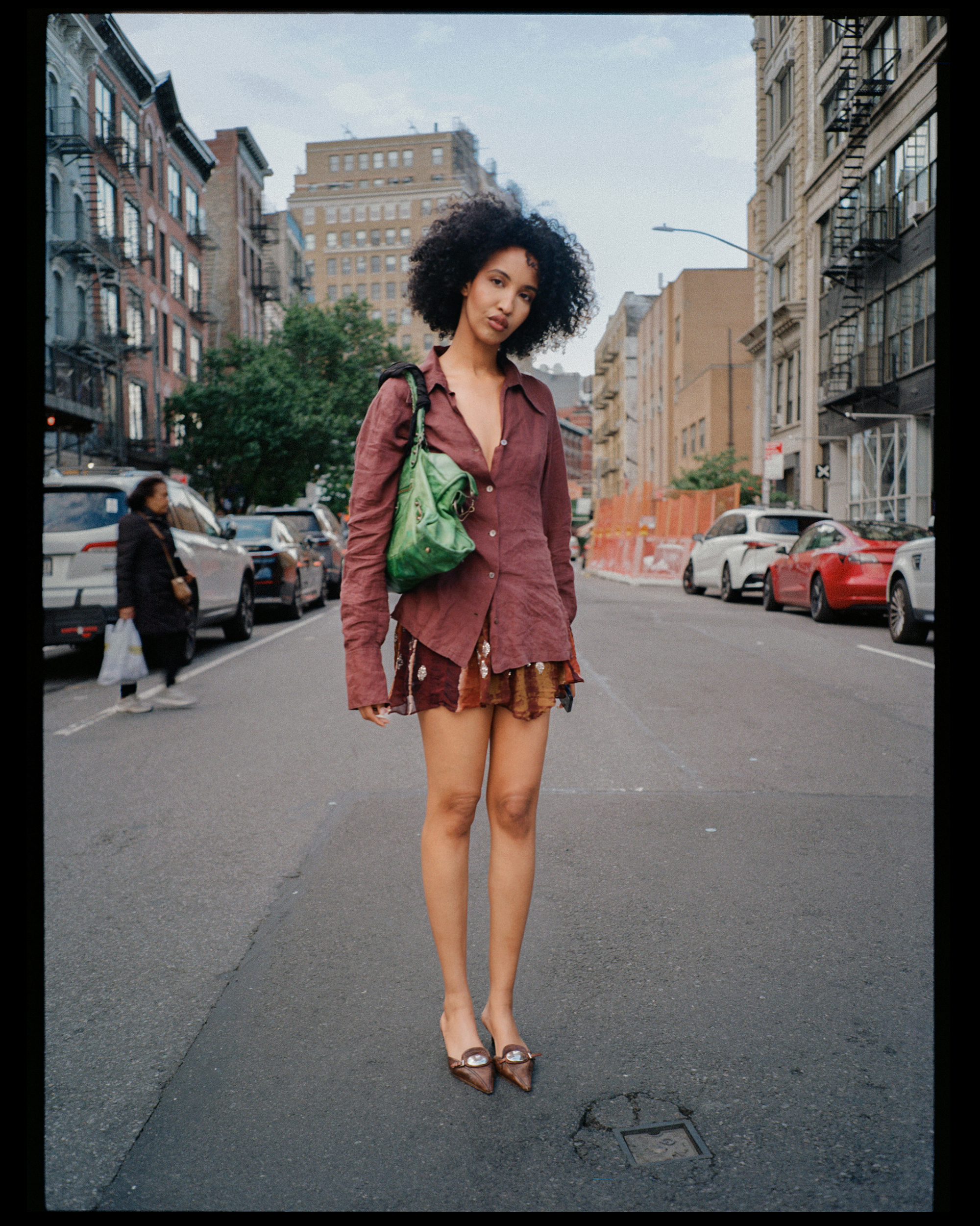 A woman with curly hair standing barefoot on a city street, wearing a loose burgundy shirt, patterned shorts, and brown shoes, with parked cars and buildings in the background.