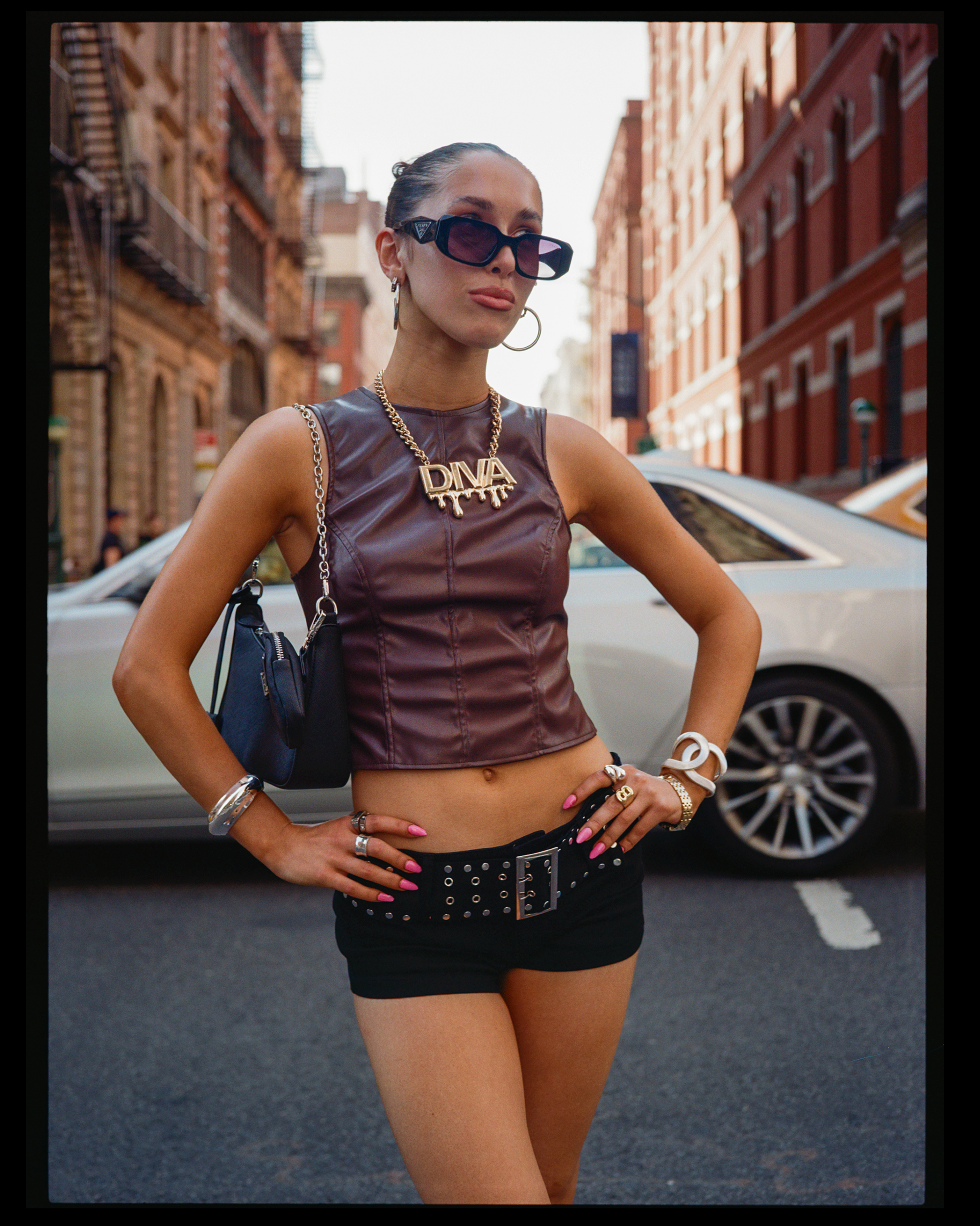 A woman standing on a city street wearing oversized black sunglasses, a sleeveless leather top, and a large gold necklace with the name 'DIVA.' She is also wearing rings, bracelets, and a belt, with her hand resting on her hip. In the background, the
