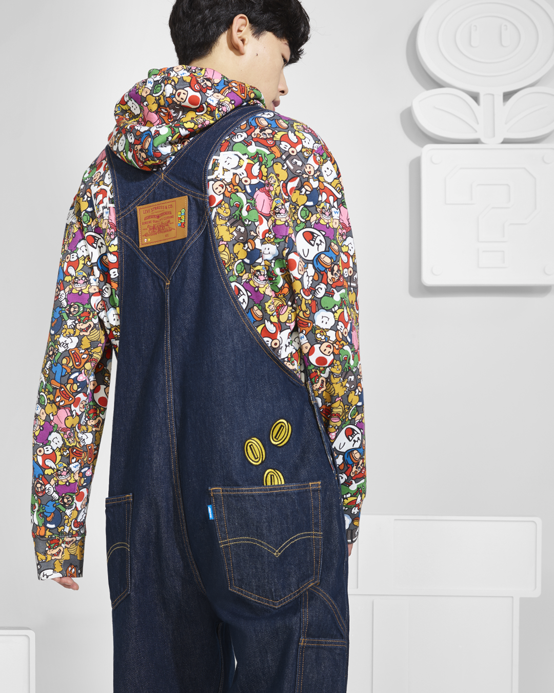 A young man wearing a colorful Mario-themed hoodie and denim overalls, standing in front of white wall art featuring question marks and a smiling face with closed eyes.