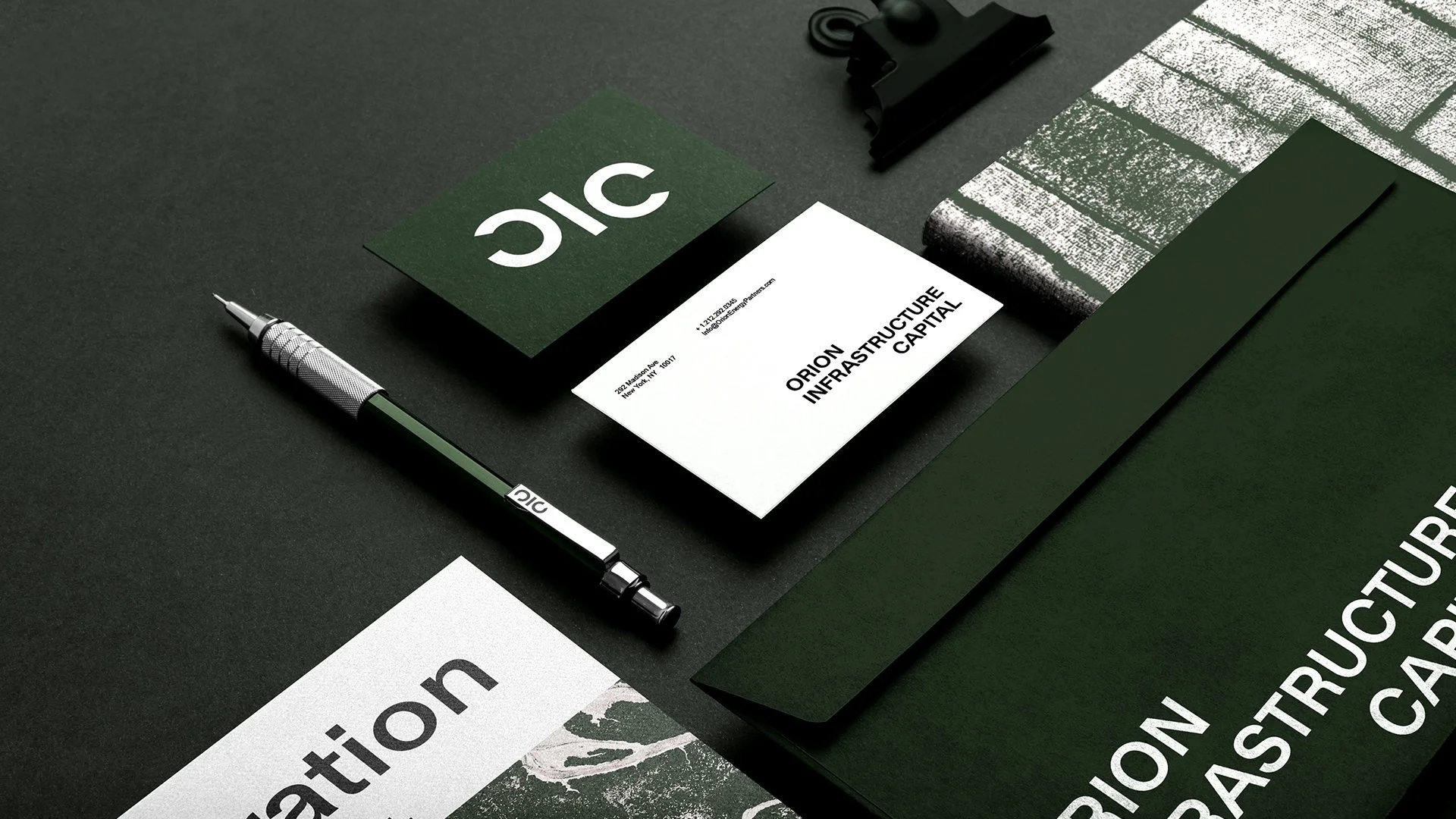 A dark green and white business presentation or branding setup on a black surface, including a pen, business cards reading "ORION INFRASTRUCTURE CAPITAL," a black binder clip, textured and printed materials, and green folders.