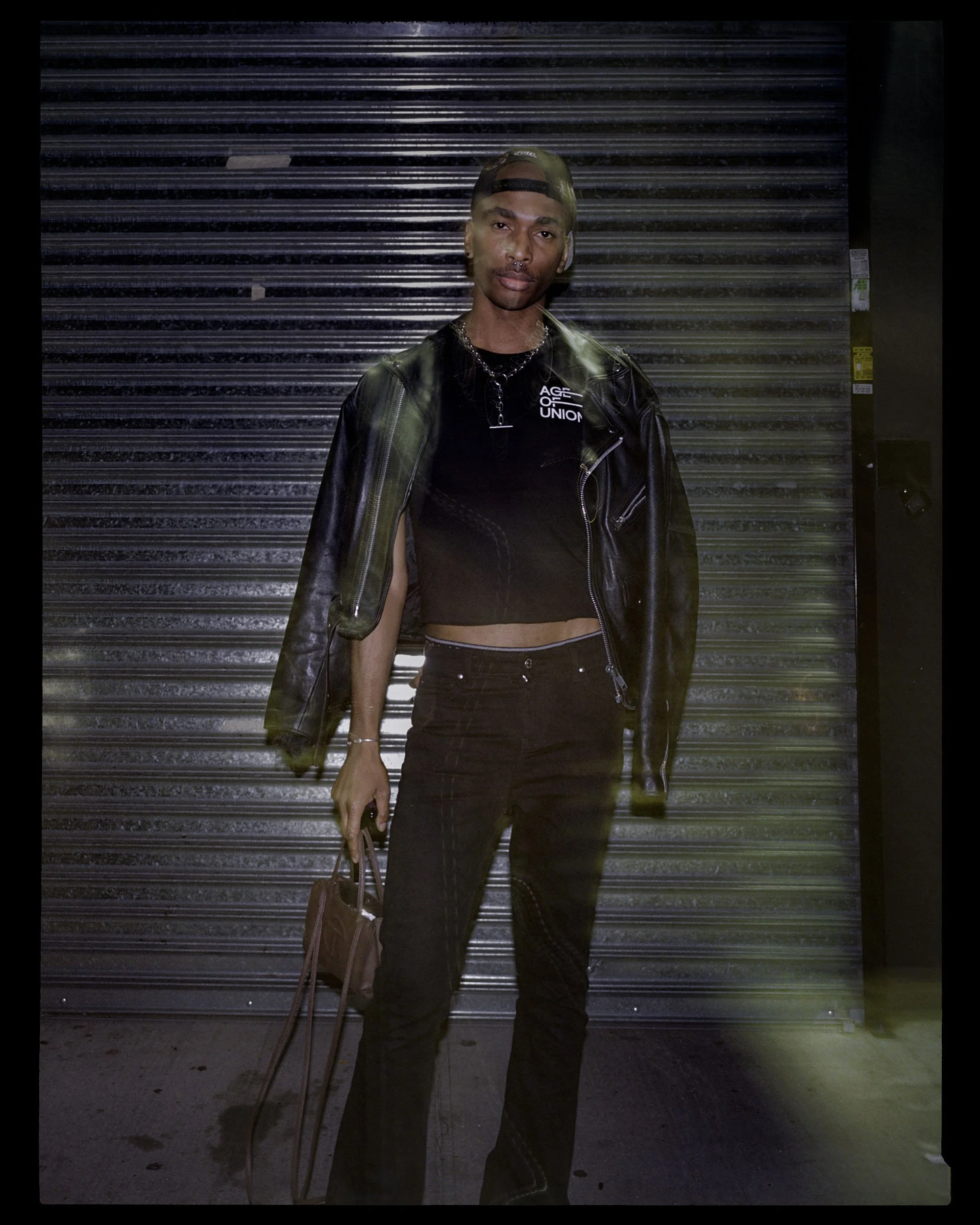 Person standing in front of metal roll-up door at night, wearing a black leather jacket, black crop top, and black pants, holding a brown bag, with a cap worn backwards.