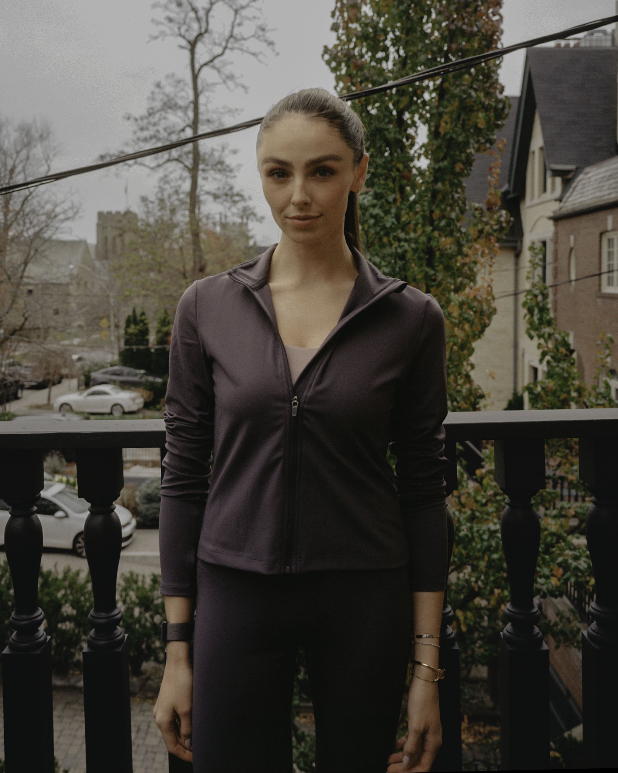 A young woman with dark hair tied back in a ponytail stands outdoors on a cloudy day. She is wearing a dark purple zip-up athletic jacket and black pants, with gold bracelets and a watch on her left wrist. Behind her, there are leafless trees, a few houses, and a cloudy sky.