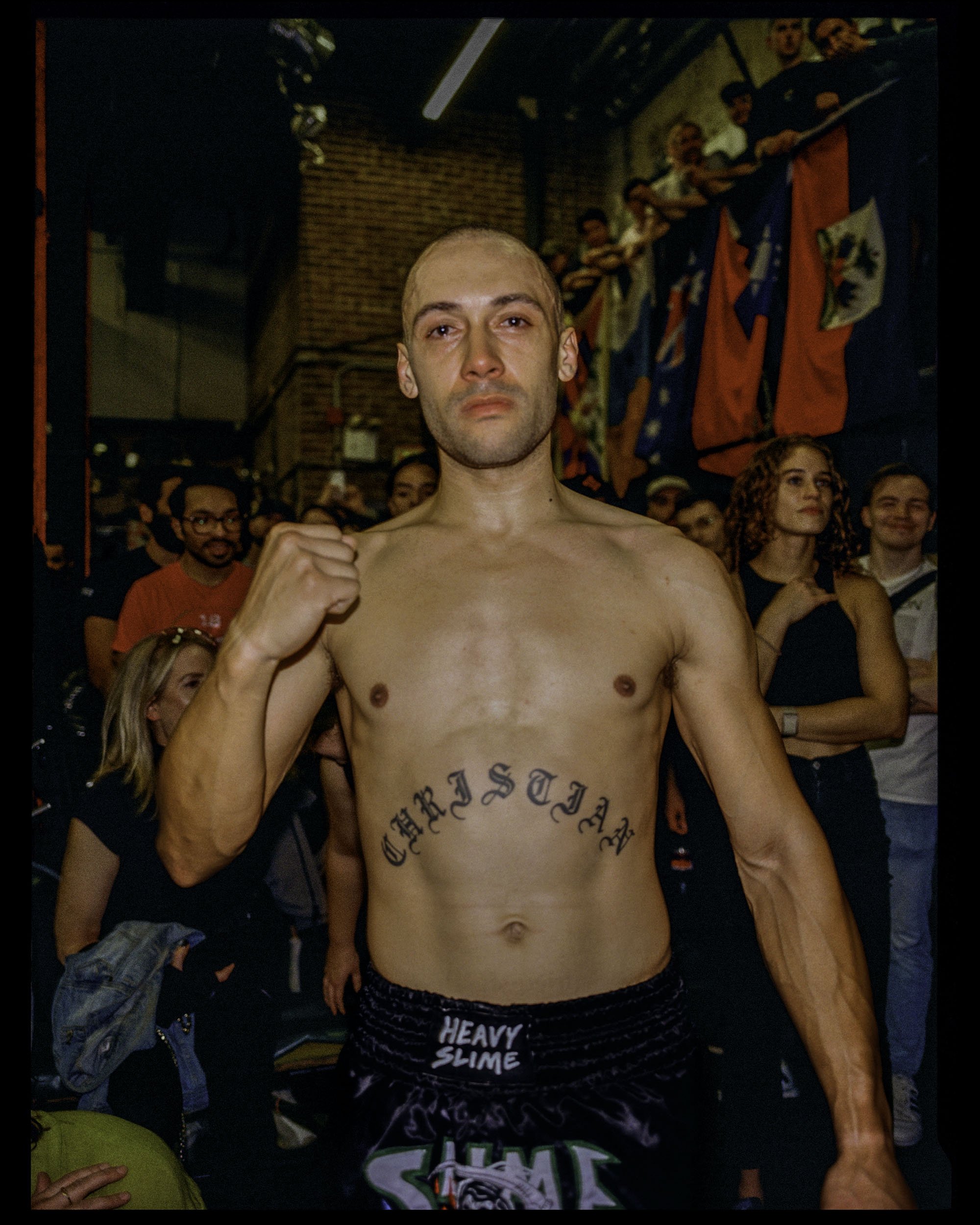 A shirtless man with a shaved head and tattoos on his chest and abdomen, standing in a crowd at an indoor event. Behind him are several people, some smiling, and colorful flags hanging from the ceiling.