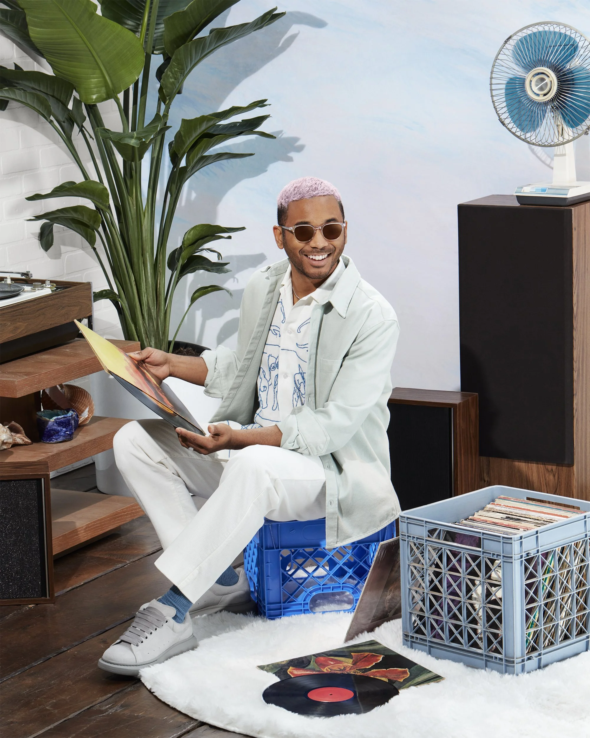 A man with short, light pink hair, sunglasses, and a white shirt with a patterned design, sitting on a blue crate, reading a vinyl record sleeve. He is surrounded by vintage decor including a record player, vinyl records, large green plant, and a blue and white fan in a cozy, retro-styled room.