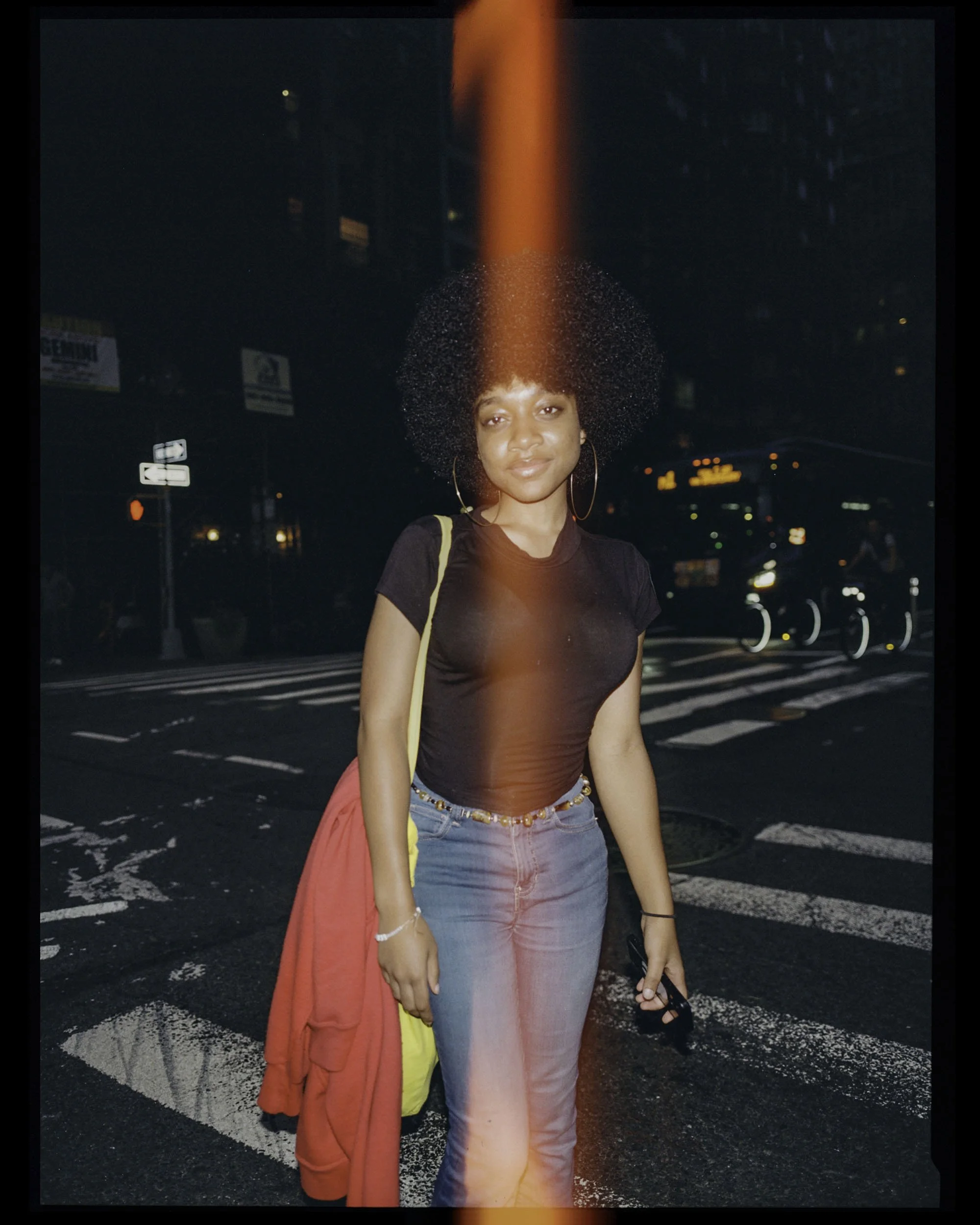 A young woman with curly hair and hoop earrings standing on a city street at night, wearing a black shirt, light-colored jeans, with a red and yellow jacket tied around her waist, holding a black phone in her right hand.