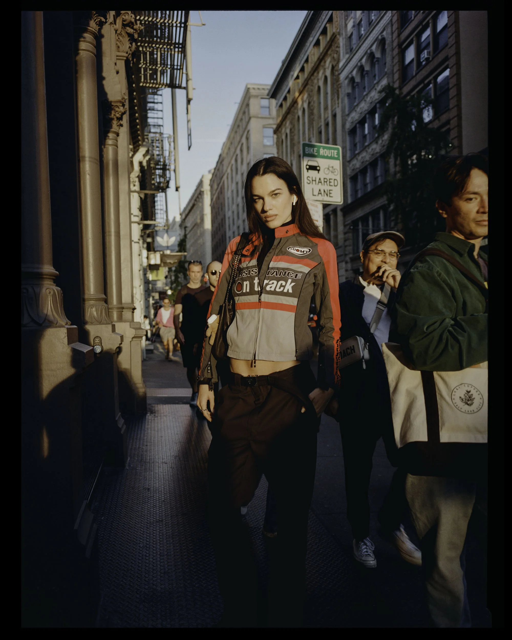 A young woman walking on a city street during daylight, wearing a racing-style jacket with 