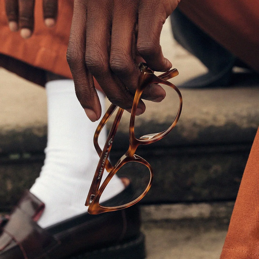 Close-up of an adult's hand holding a pair of tortoiseshell eyeglasses with the brand name 'Warby Parker' visible on the temple, with a person's legs wearing white socks and dark shoes in the background.