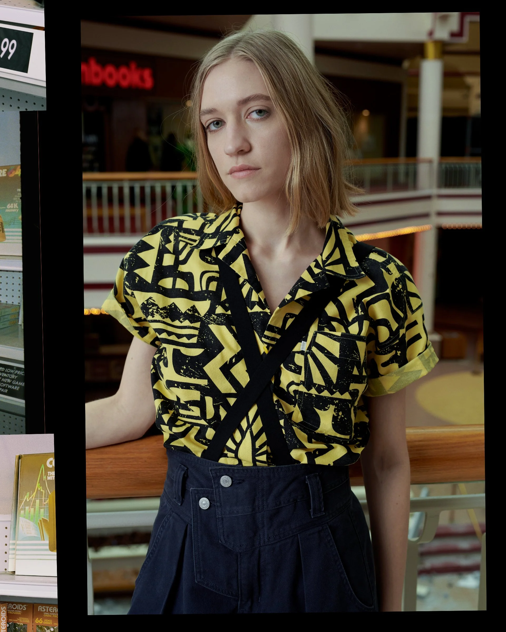 A young woman with light skin and shoulder-length blond hair, wearing a yellow and black patterned shirt and black high-waisted pants, standing inside a store near shelves of books or magazines.