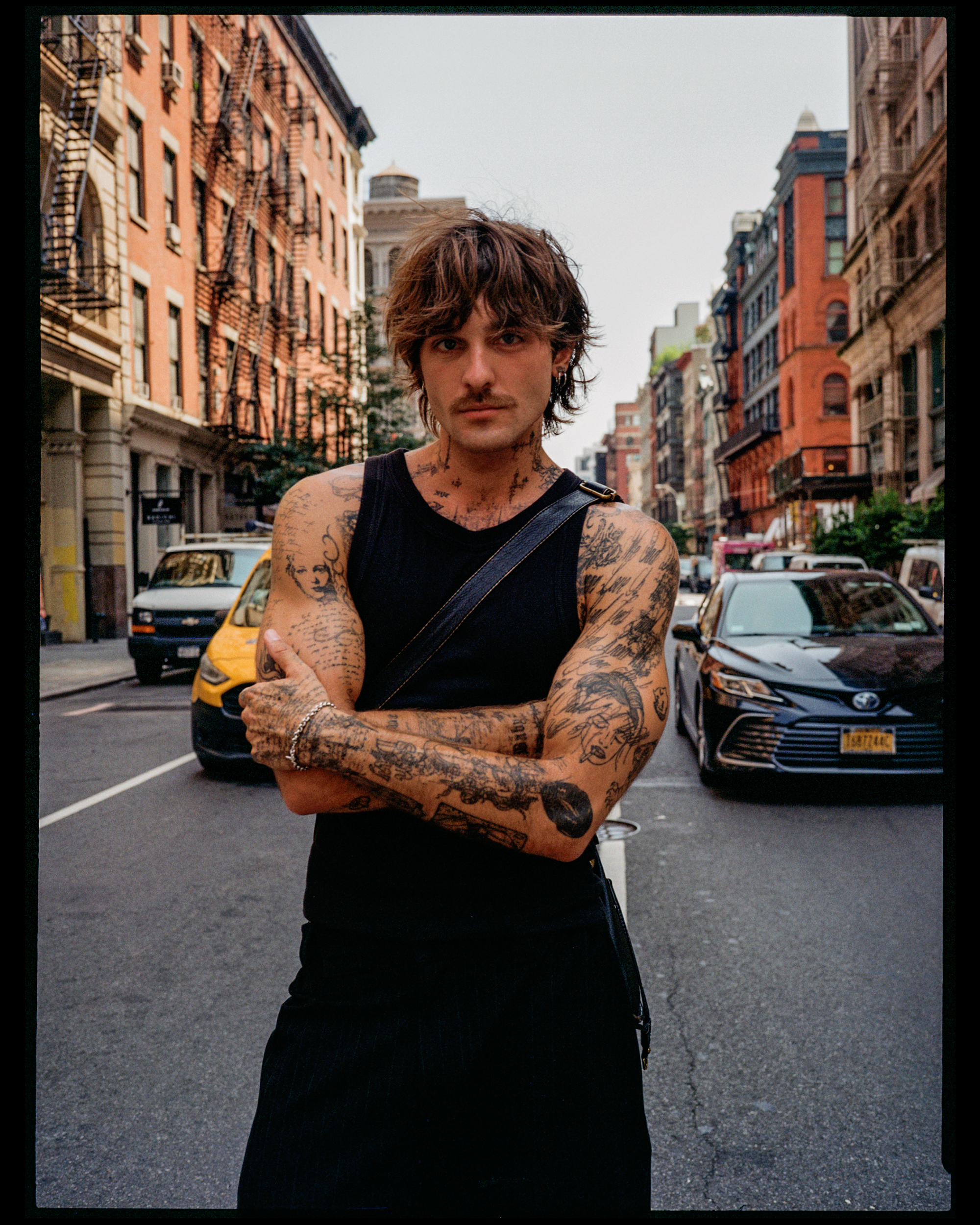 A young man with tattoos and earrings in an urban street scene with parked cars and multistory brick buildings.