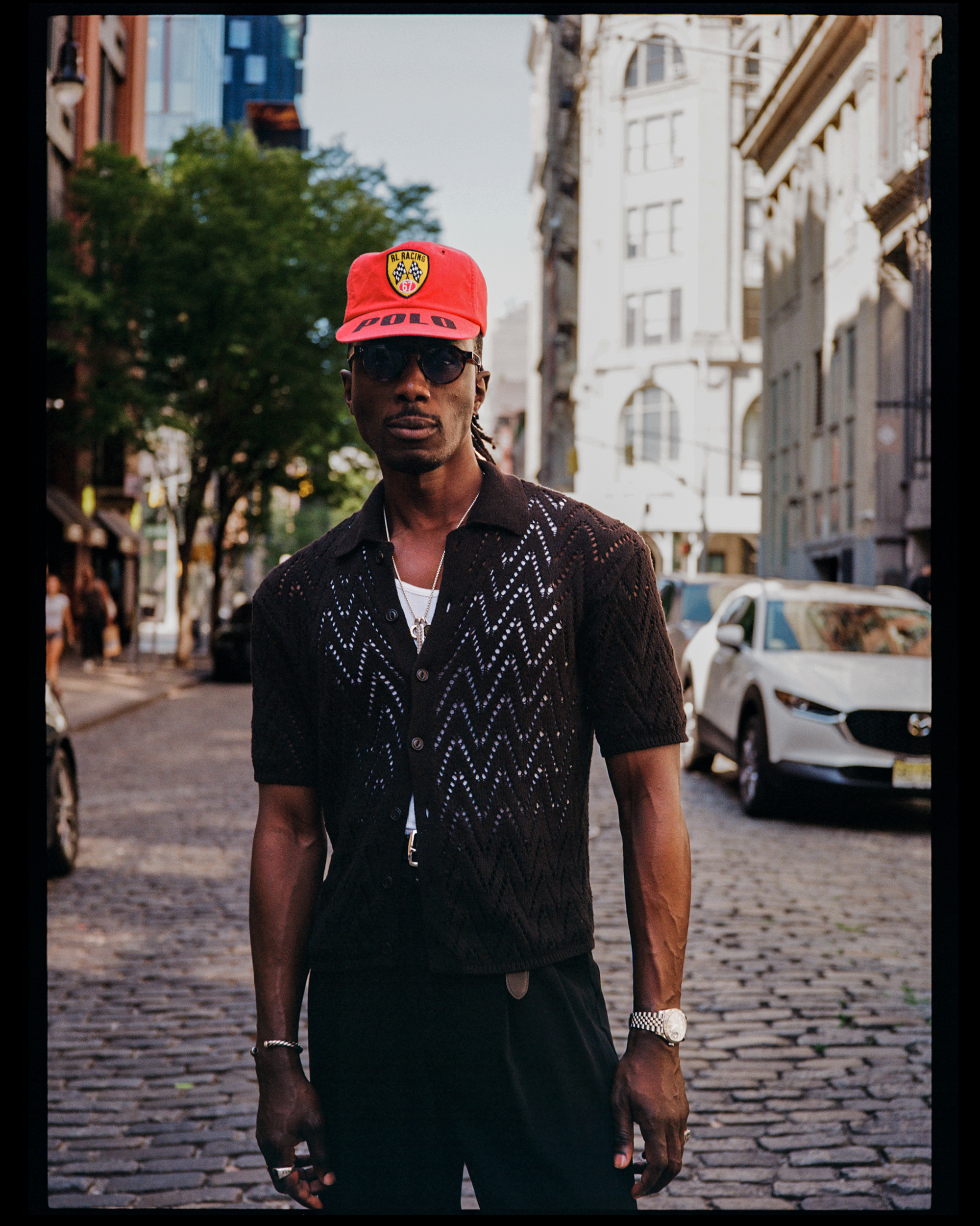 Man walking on cobblestone street in urban area, wearing a red cap with a racing logo, sunglasses, a black patterned short-sleeve shirt, and jewelry, with city buildings and parked cars in the background.