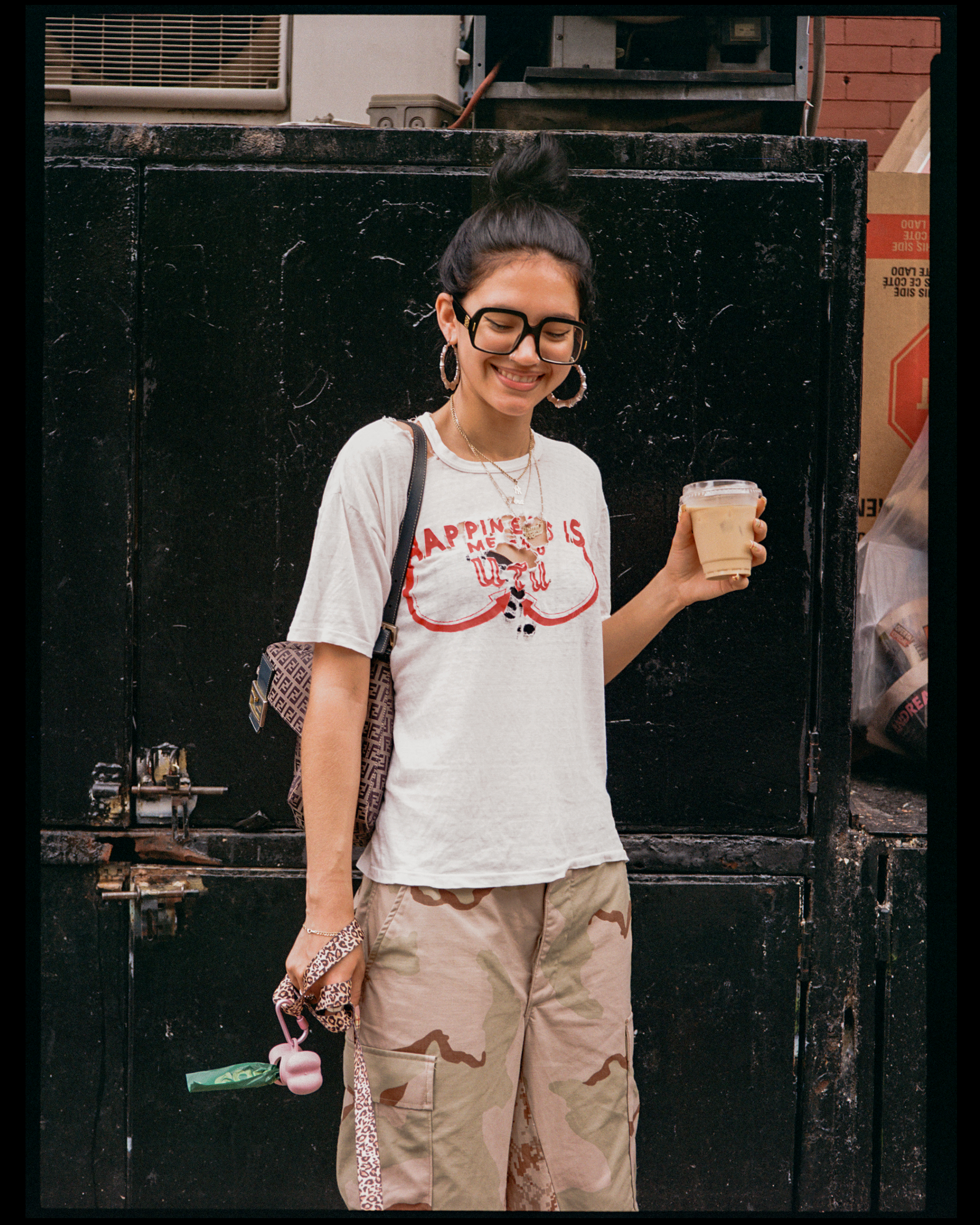 A woman with dark hair styled in a top bun, wearing large glasses, earrings, and a white T-shirt with red text, smiles while holding a coffee cup.