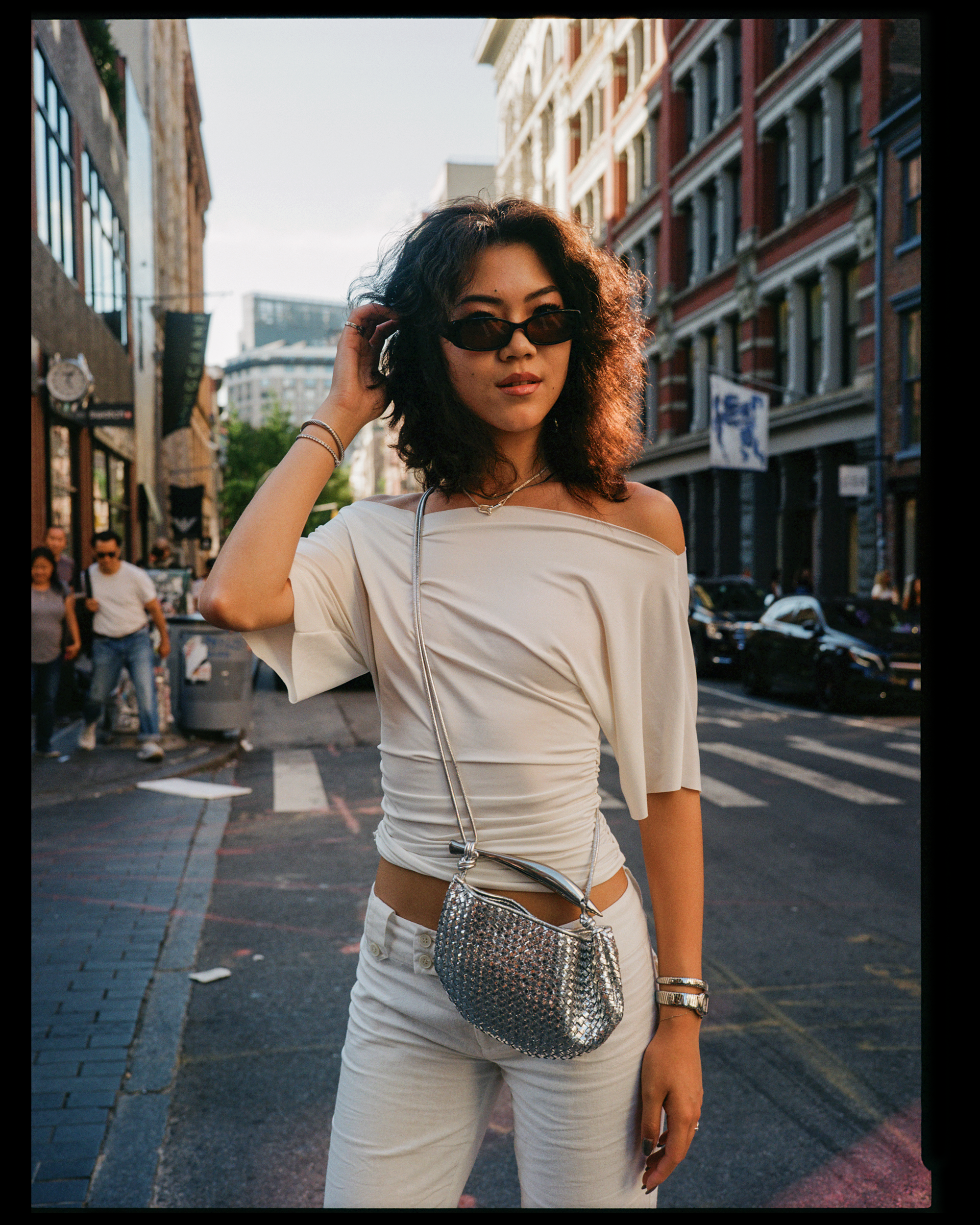 A woman with curly dark hair wearing sunglasses, a white off-shoulder top, and white pants in an urban street setting with buildings and people in the background.