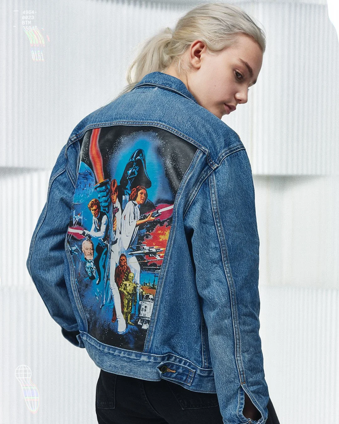 A young woman with blonde hair in a ponytail wearing a denim jacket with a Star Wars graphic on the back, standing sideways in a bright, modern room.