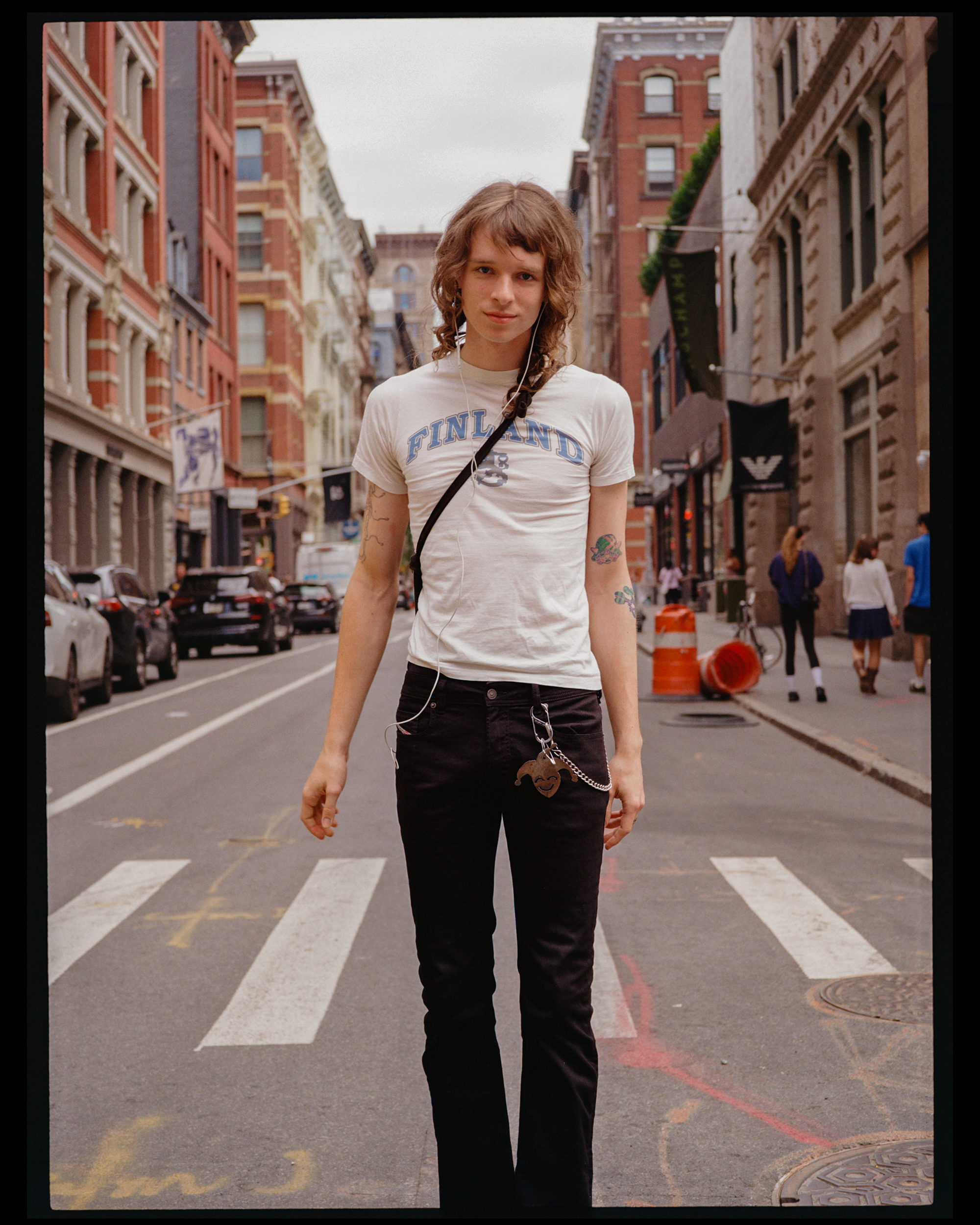 A young woman with curly brown hair walking down a city street. She is wearing a white T-shirt with the word "Finland" printed on it and black pants. She has colorful tattoos on her arms and is listening to earphones.