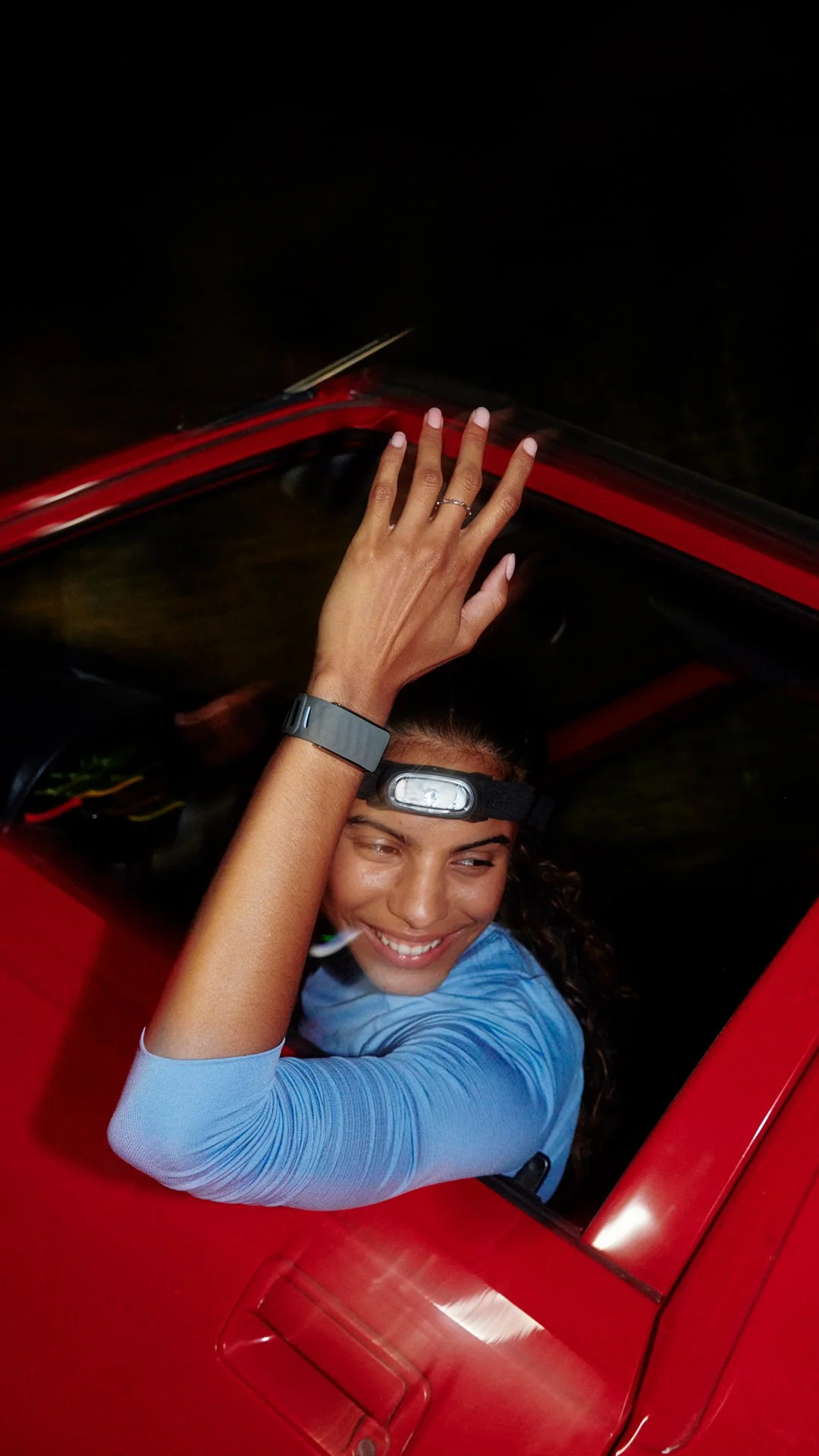 Smiling woman with headlamp and fitness tracker smiling out of a red vehicle's window at night