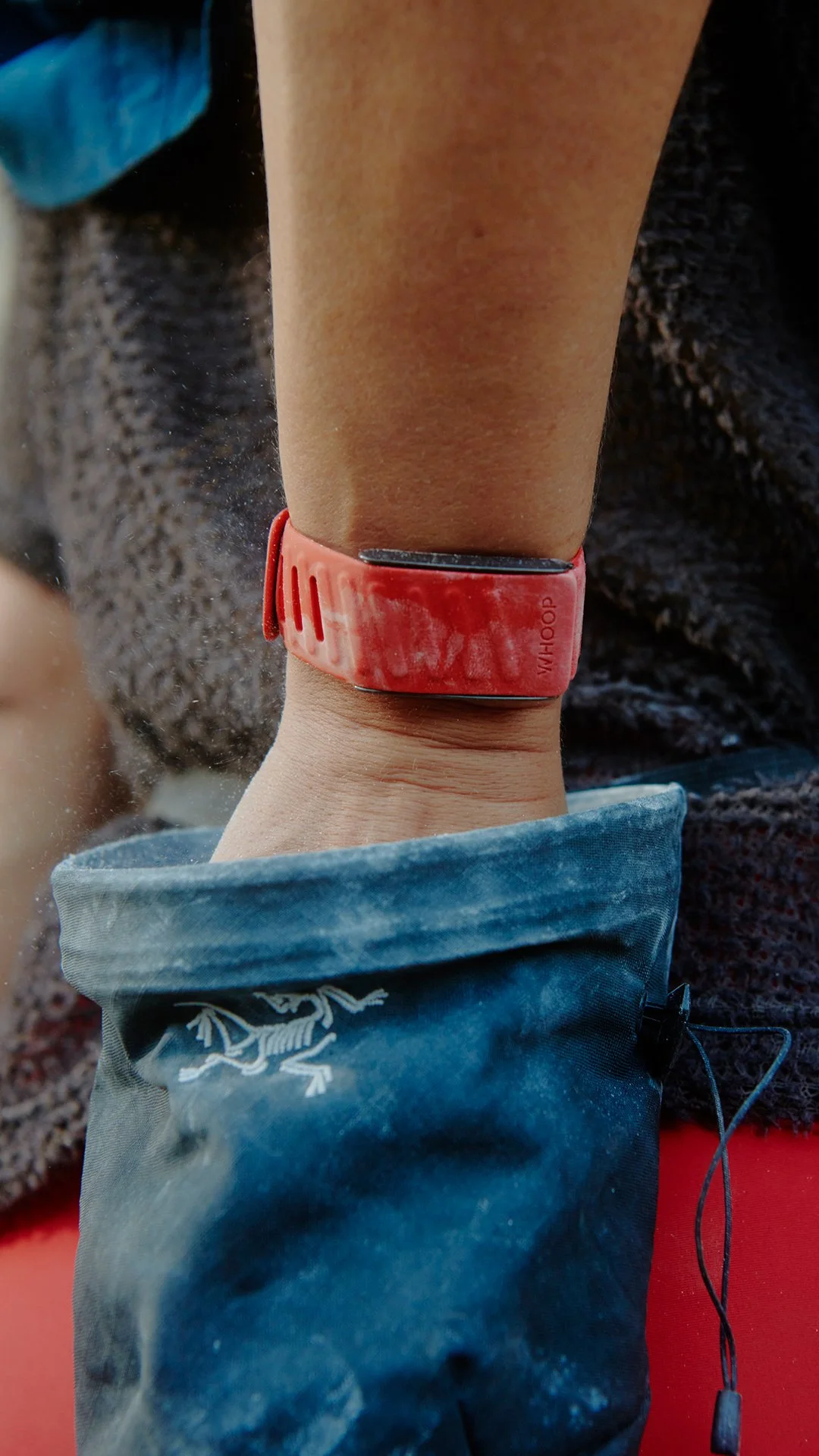 Close-up of a person's wrist wearing a red activity tracker or smartwatch. The person is dressed in outdoor gear, with a blue sleeve and a greenish jacket, indicating they are engaging in outdoor activity, likely hiking or trail running.