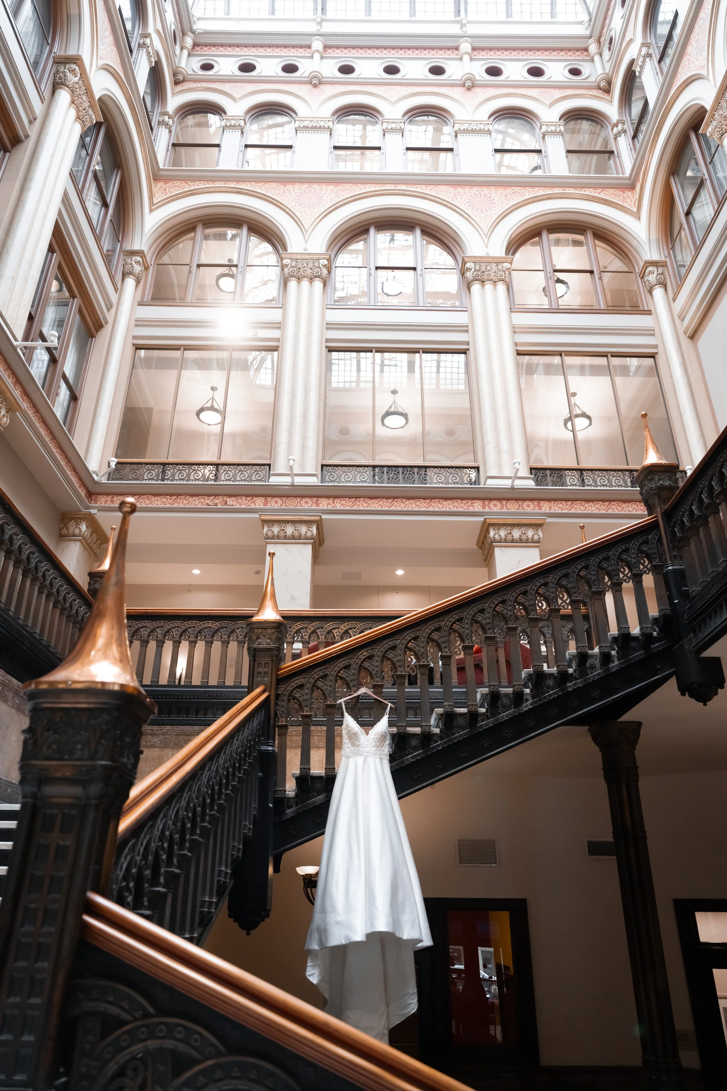 Elegant interior of a building with a staircase, large arched windows, a chandelier, and a wedding dress hanging from the staircase railing.