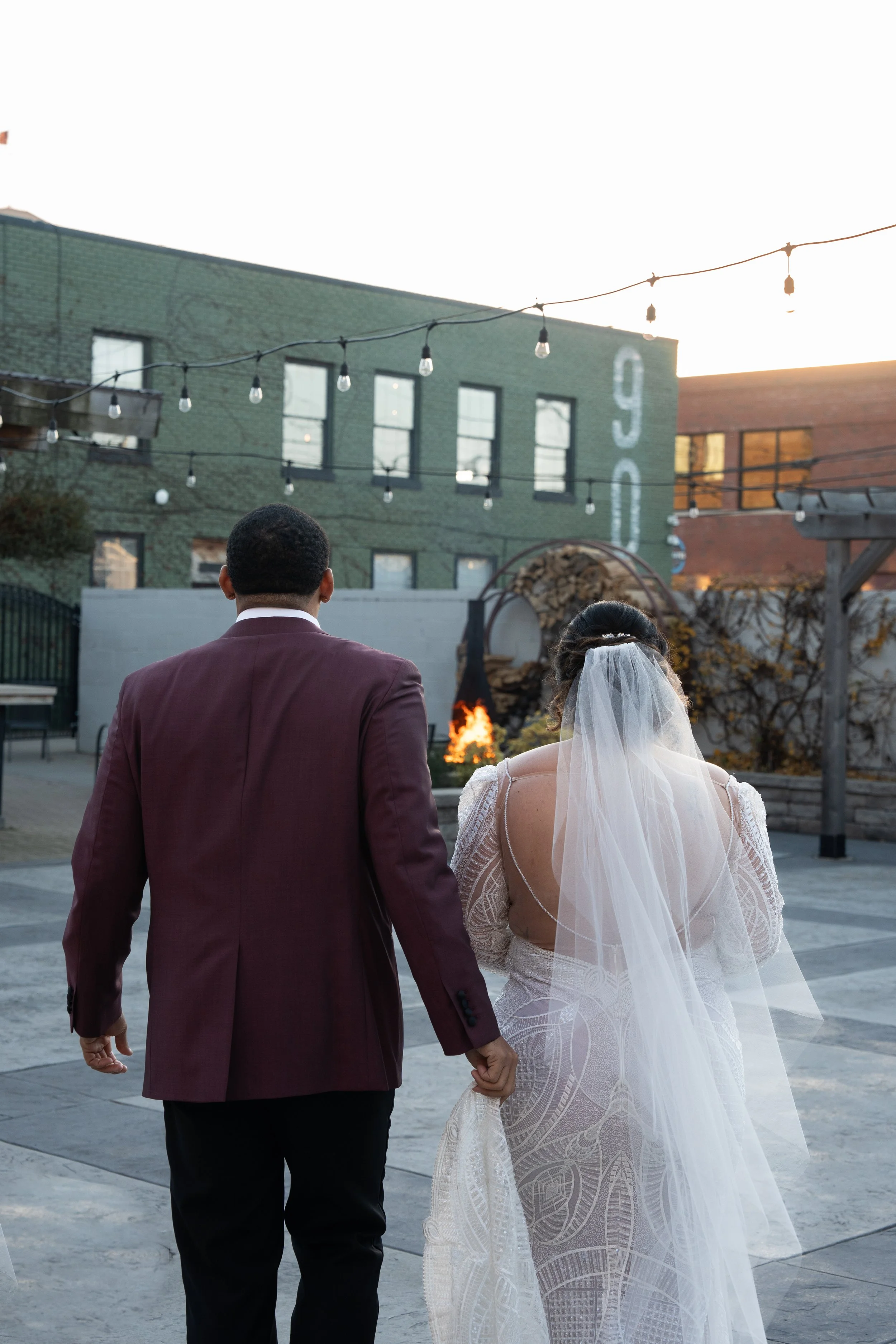 A bride and groom holding hands and walking outdoors in an urban setting during sunset, with string lights overhead and buildings in the background.