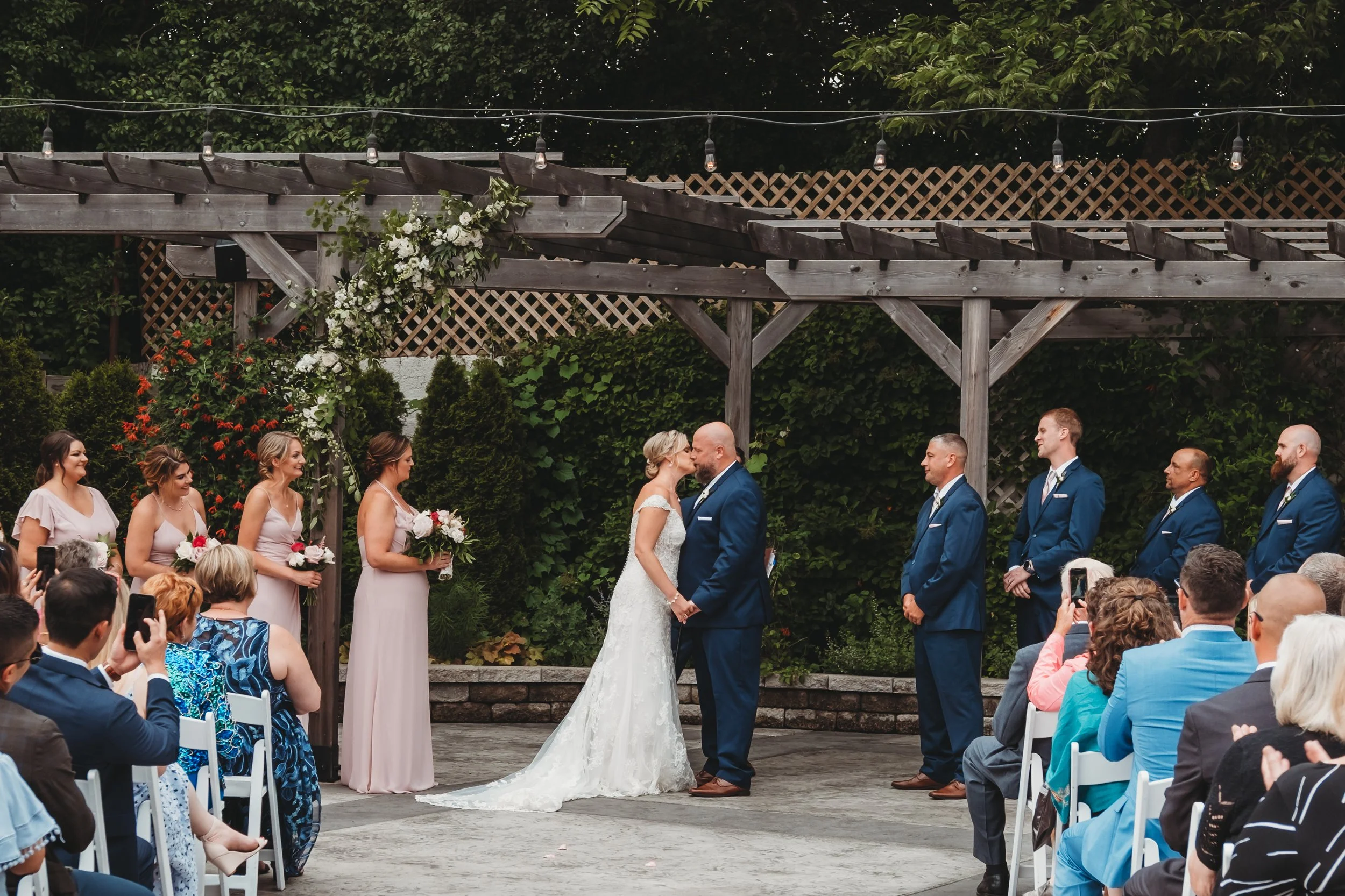 A wedding ceremony outdoors with the bride and groom kissing, surrounded by bridesmaids and groomsmen, under a wooden pergola decorated with flowers and greenery. Guests are seated and watching, some taking photos. The background has trees and a latt