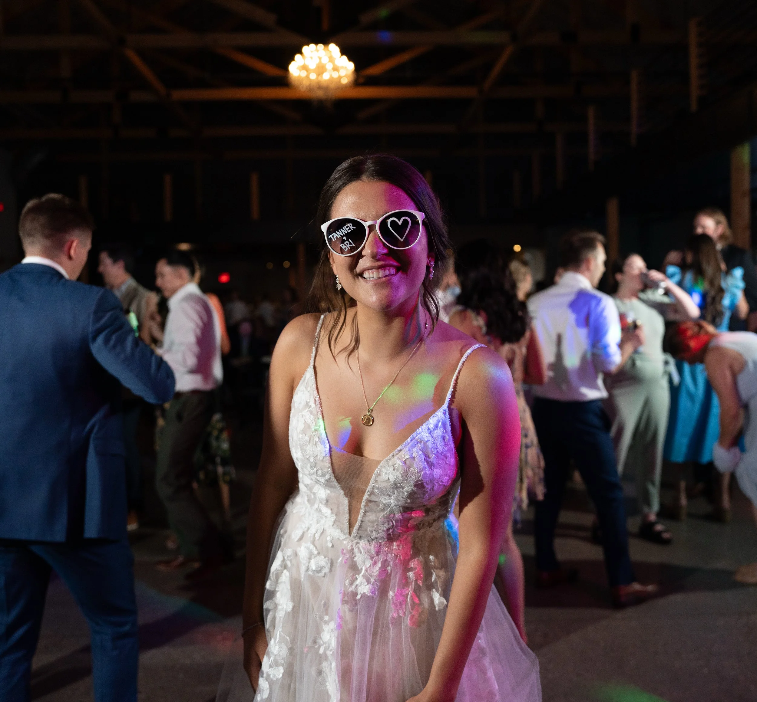 Woman wearing decorative sunglasses smiling at a party with people in the background, dim lighting, and a chandelier.