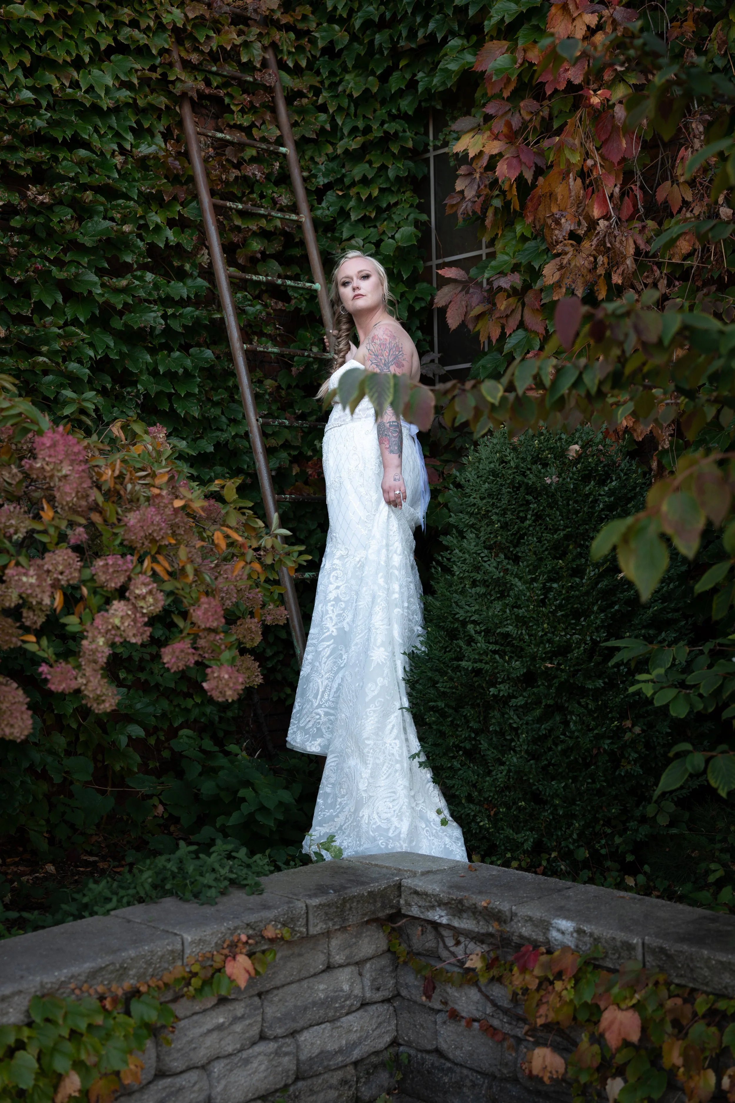 A woman in a white dress with tattoos on her arms, standing between green and reddish autumn leaves, leaning on an old, rusty ladder surrounded by foliage.