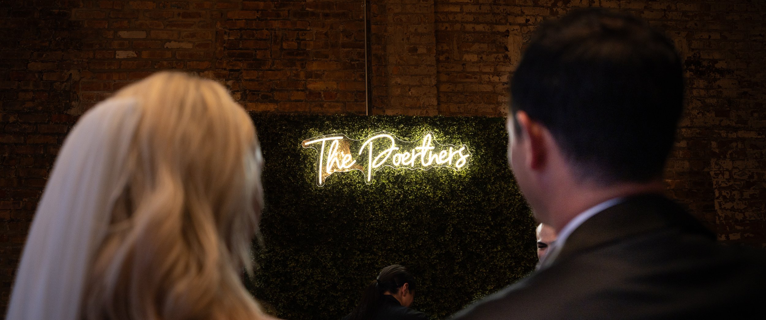 People looking at a neon sign that says 'The Poertners' on a green hedge backdrop, with a brick wall behind them.