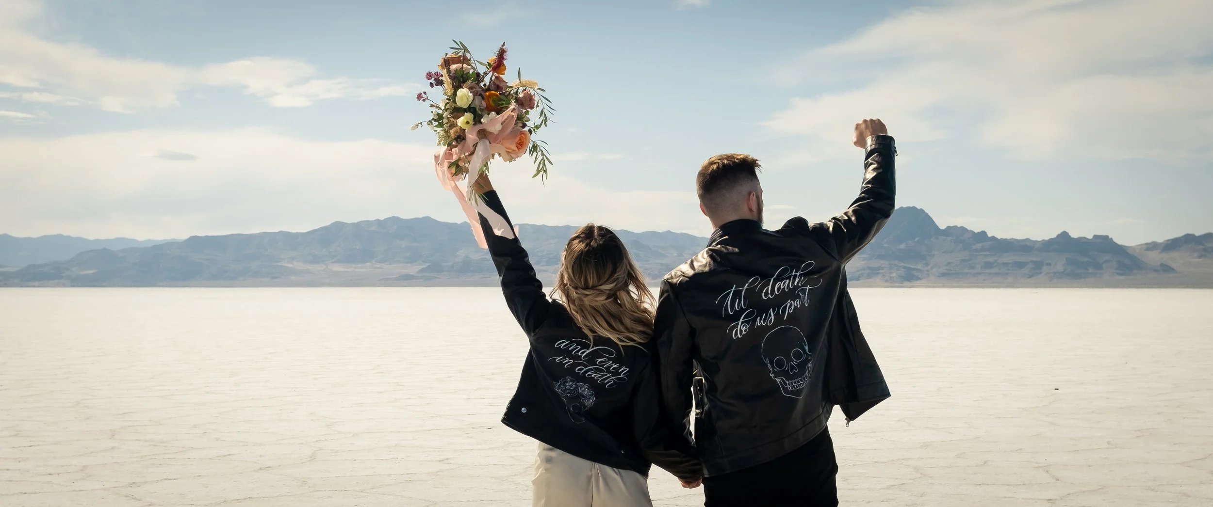 A couple holding hands by a lake shows the woman raising a bouquet of flowers, with mountains in the background, suggesting a scenic outdoor setting.