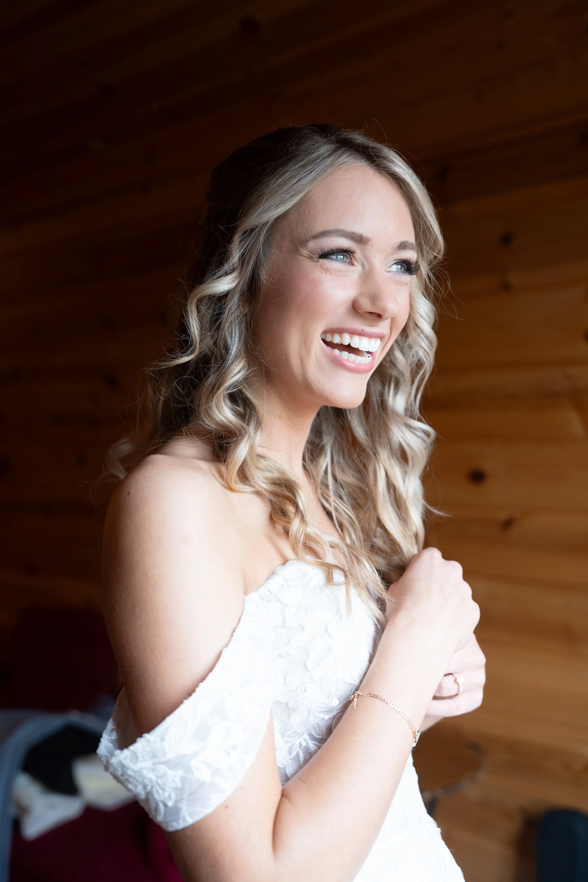 A young woman with blonde curly hair, smiling, wearing a white lace dress, standing indoors against a wooden wall.
