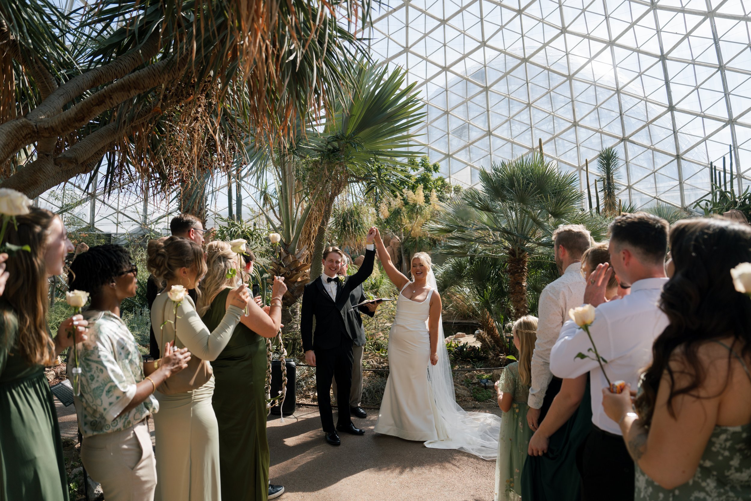 A wedding ceremony held inside a large glass conservatory, with a bride and groom standing hand in hand in front of an audience.