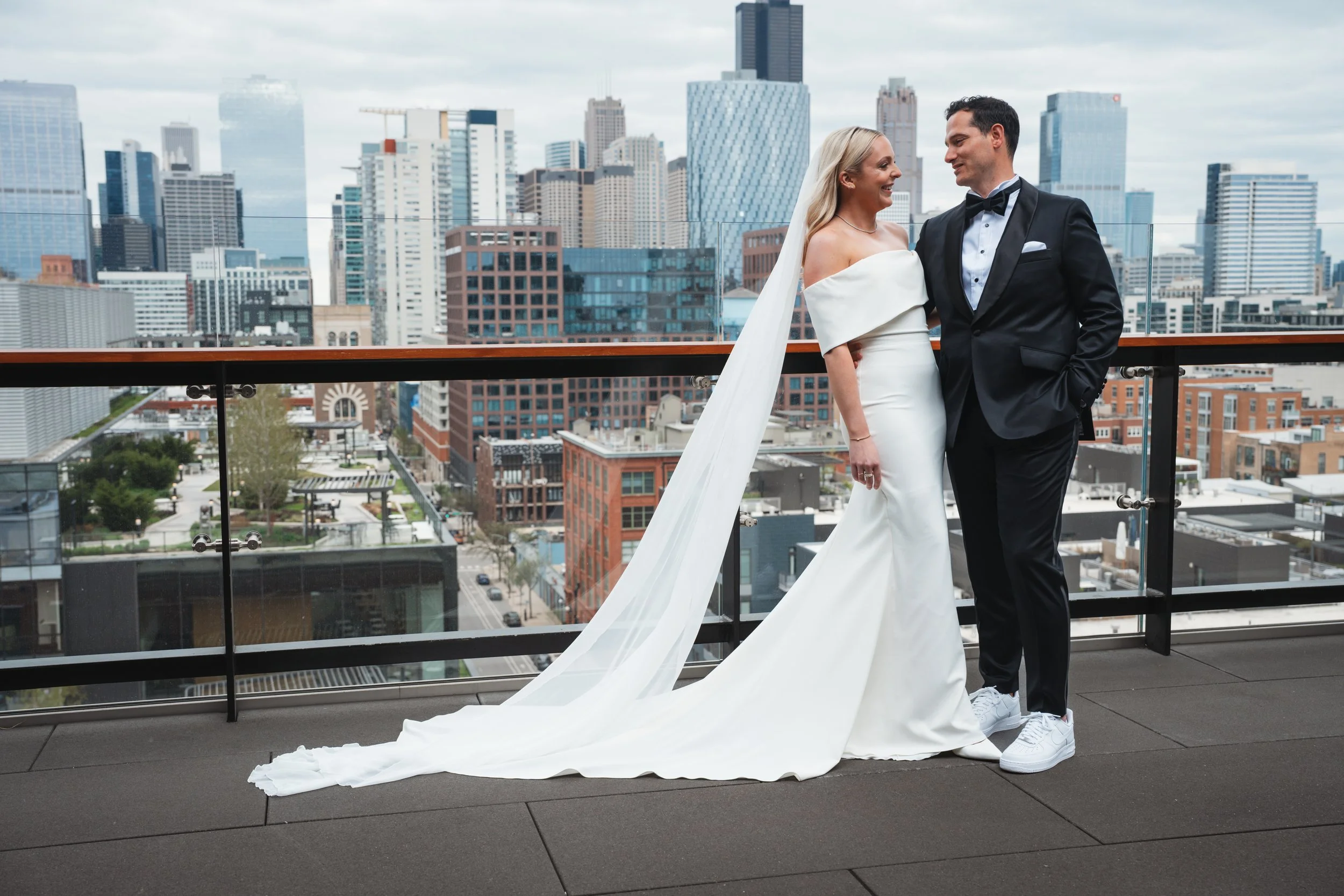 A couple in wedding attire standing on a balcony with a city skyline in the background. The bride is wearing a white off-the-shoulder wedding dress with a long train, and the groom is wearing a black tuxedo with a bow tie. They are smiling and lookin