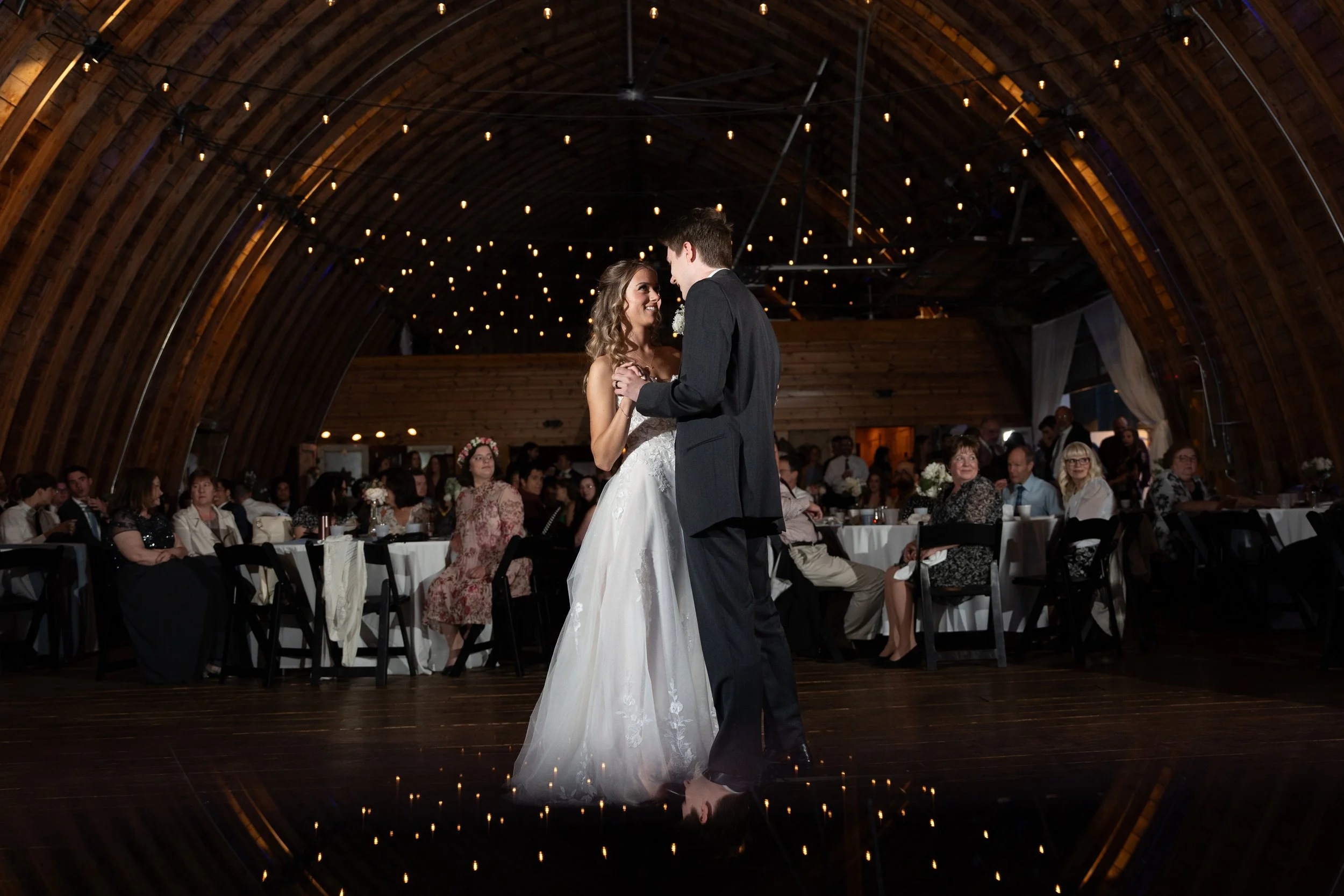 A bride and groom sharing a dance surrounded by seated guests in a rustic venue with wooden arches and string lights overhead.