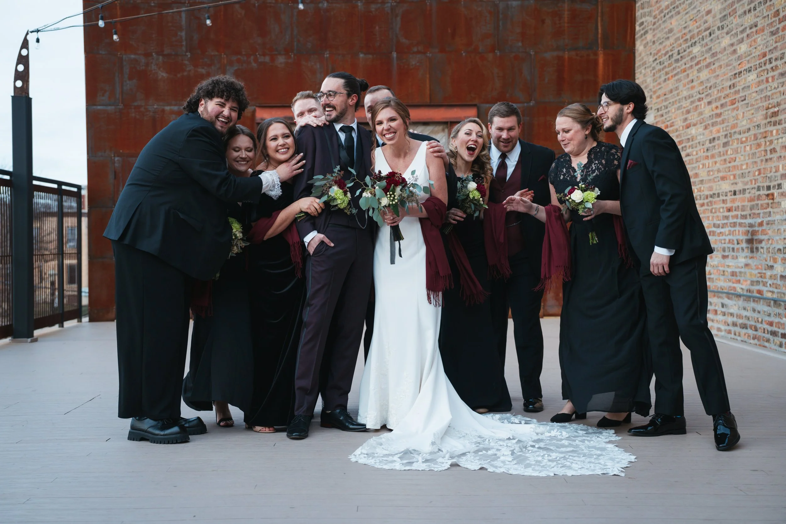 Group of people celebrating a wedding with the bride and groom at the center, all smiling and hugging each other outside on a patio with brick and rust-colored metal wall in the background.