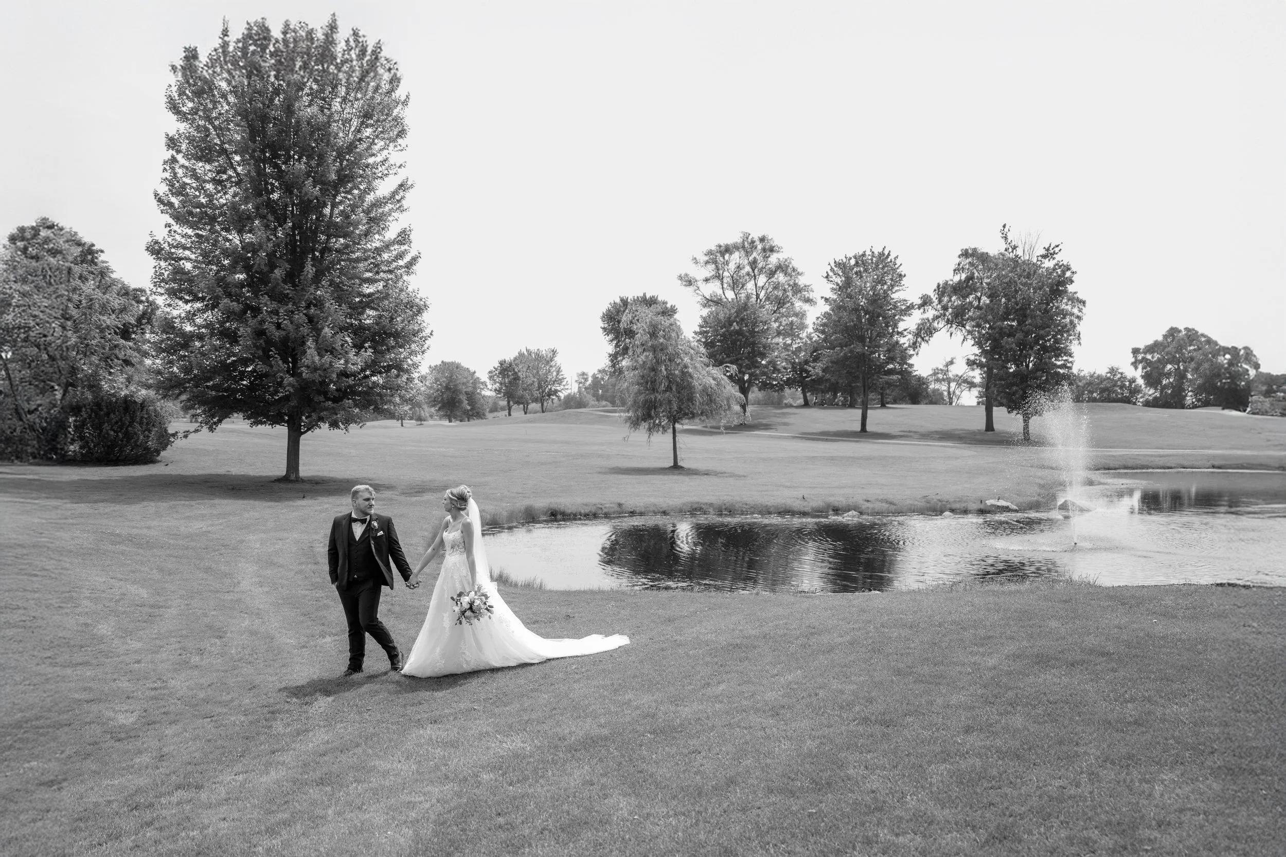 A black and white photo of a bride and groom holding hands on a grassy area near a pond, with trees and a fountain in the background.