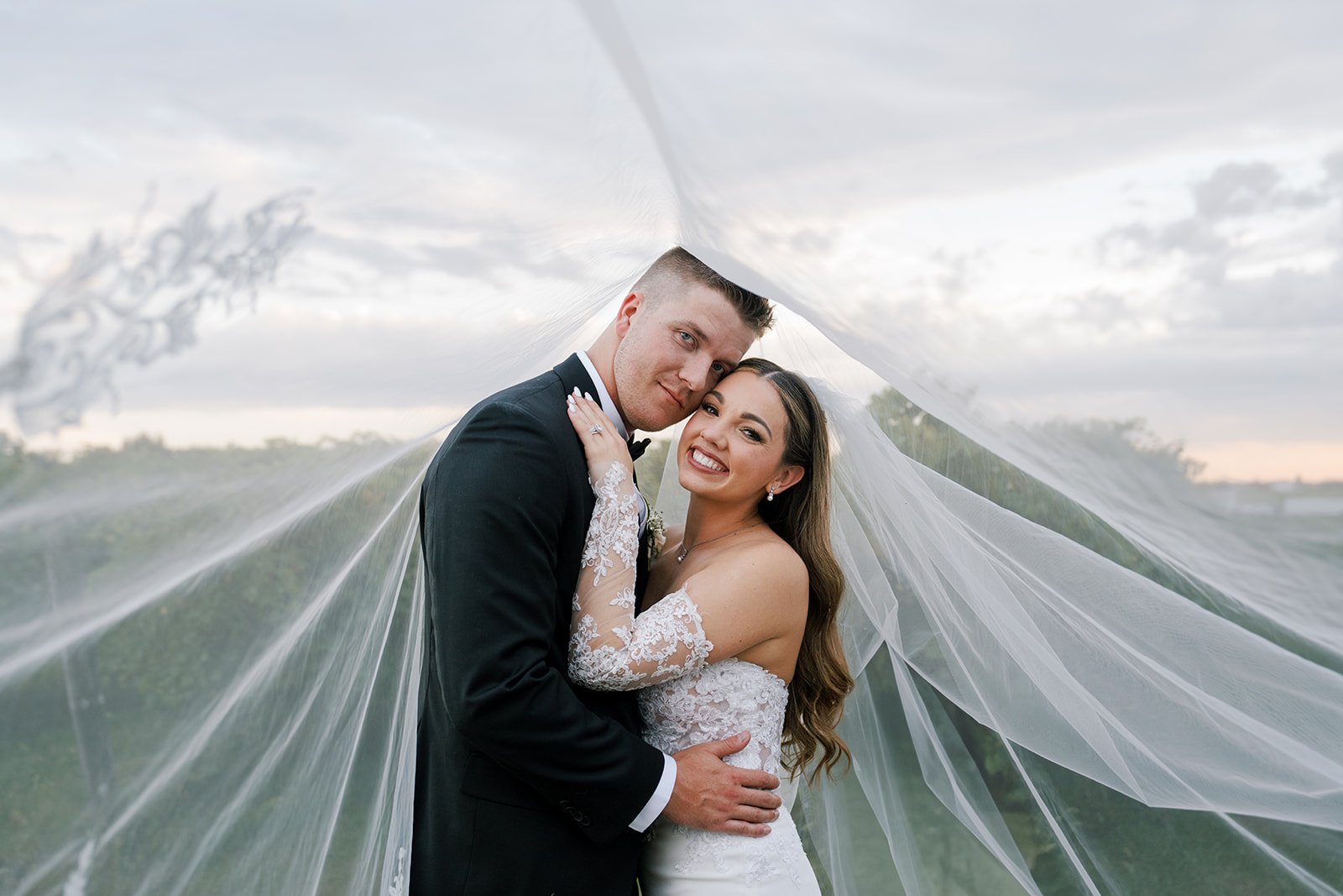 A happy bride and groom on their wedding day outdoors, with the bride's veil draped over both of them, smiling and embracing.