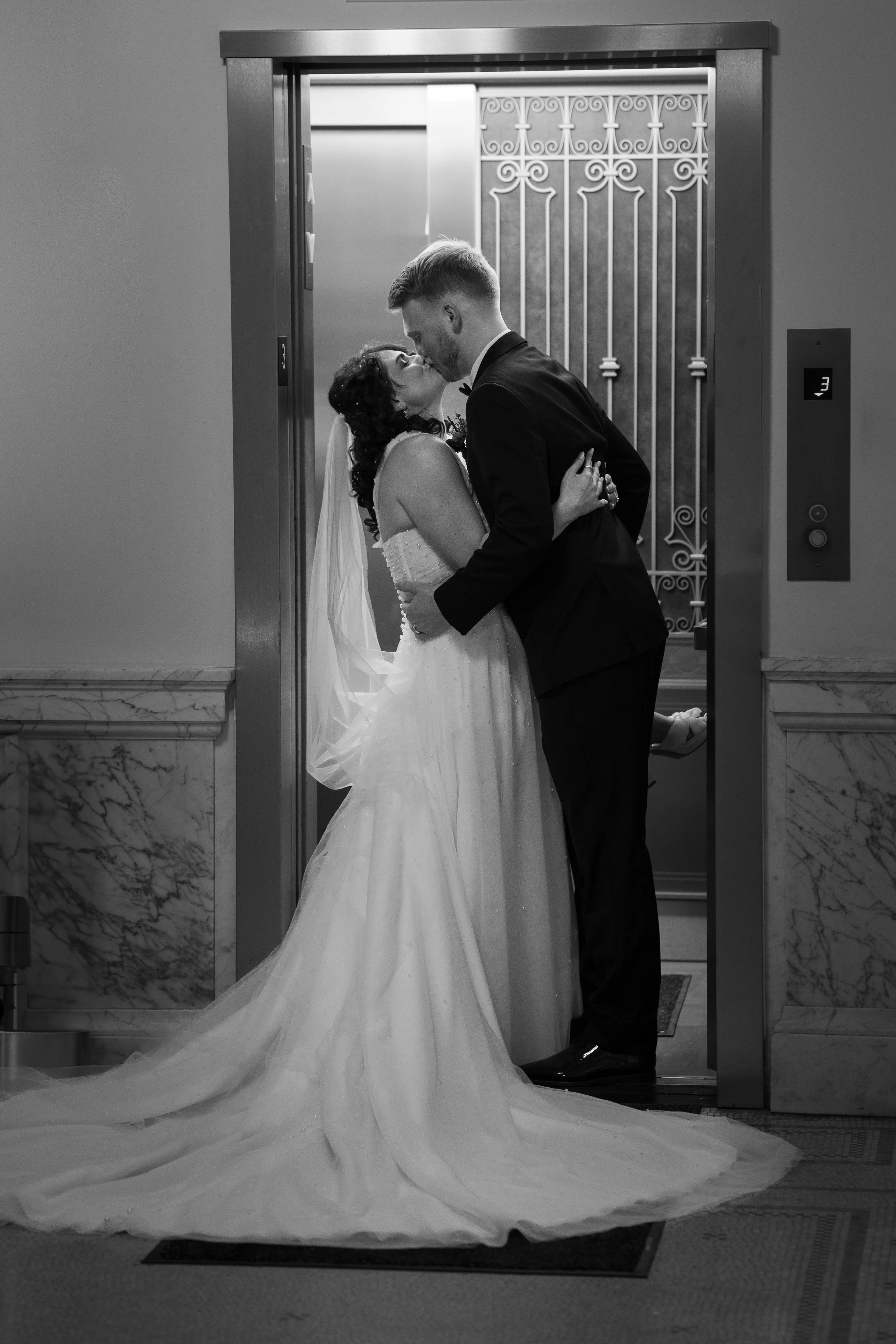 A bride and groom share a kiss in an elevator with the doors open, bride in a gown with a long train, groom in a tuxedo, both holding each other.