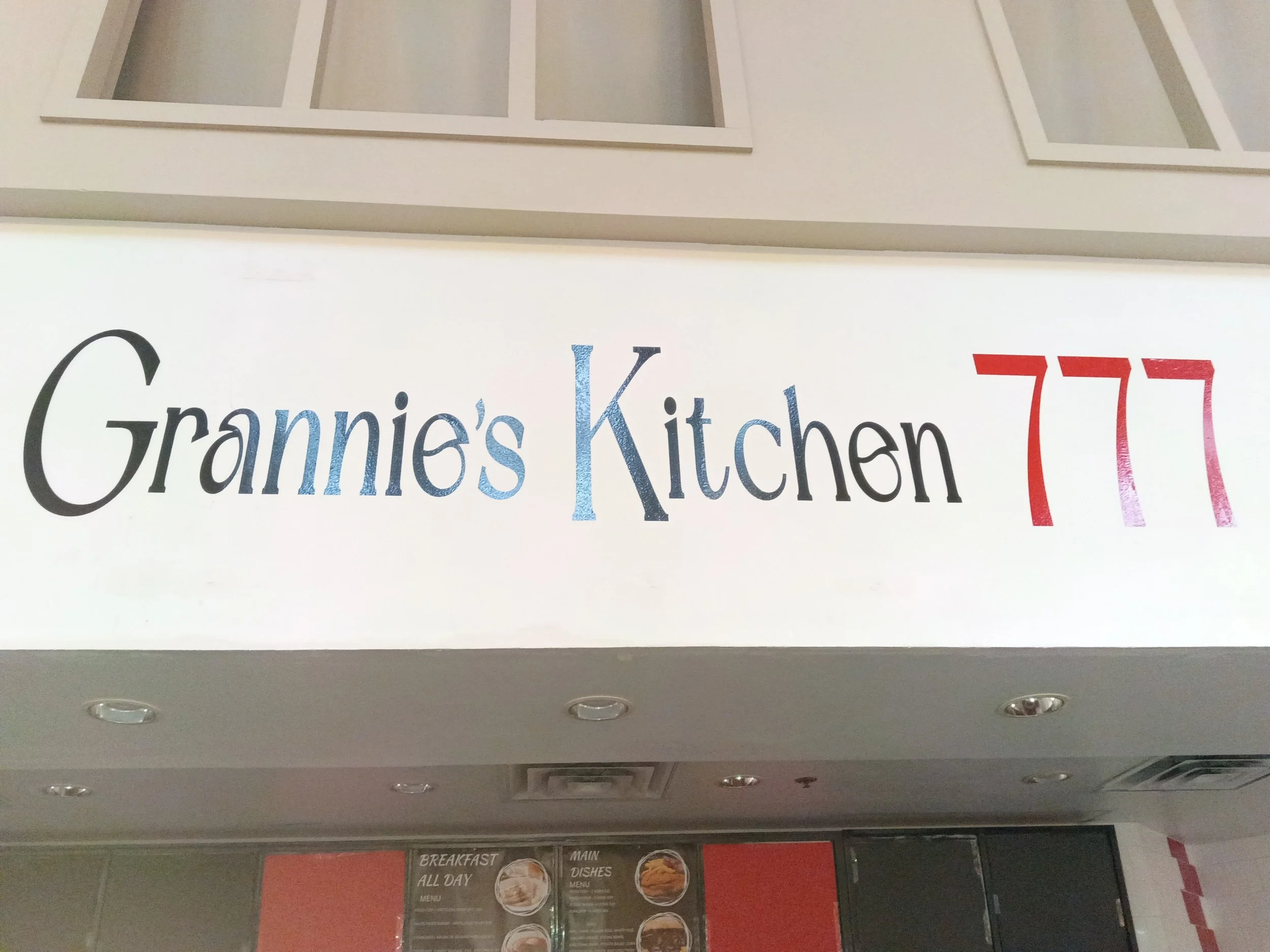 Part of a restaurant or cafe menu board beneath ceiling lights, with the words 'Grammie’s Kitchen' visible and red stylized numbers '77.'