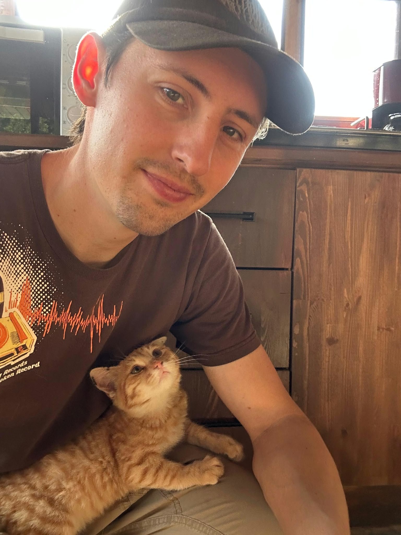Ein junger Mann mit einem schwarzen Cap, braunem T-Shirt mit einem orangefarbenen Druck und einem kleinen orangefarbenen Kätzchen, das in seinem Schoß liegt, sitzt in einem Raum mit Holzmöbeln und einer großen Fensteröffnung im Hintergrund.