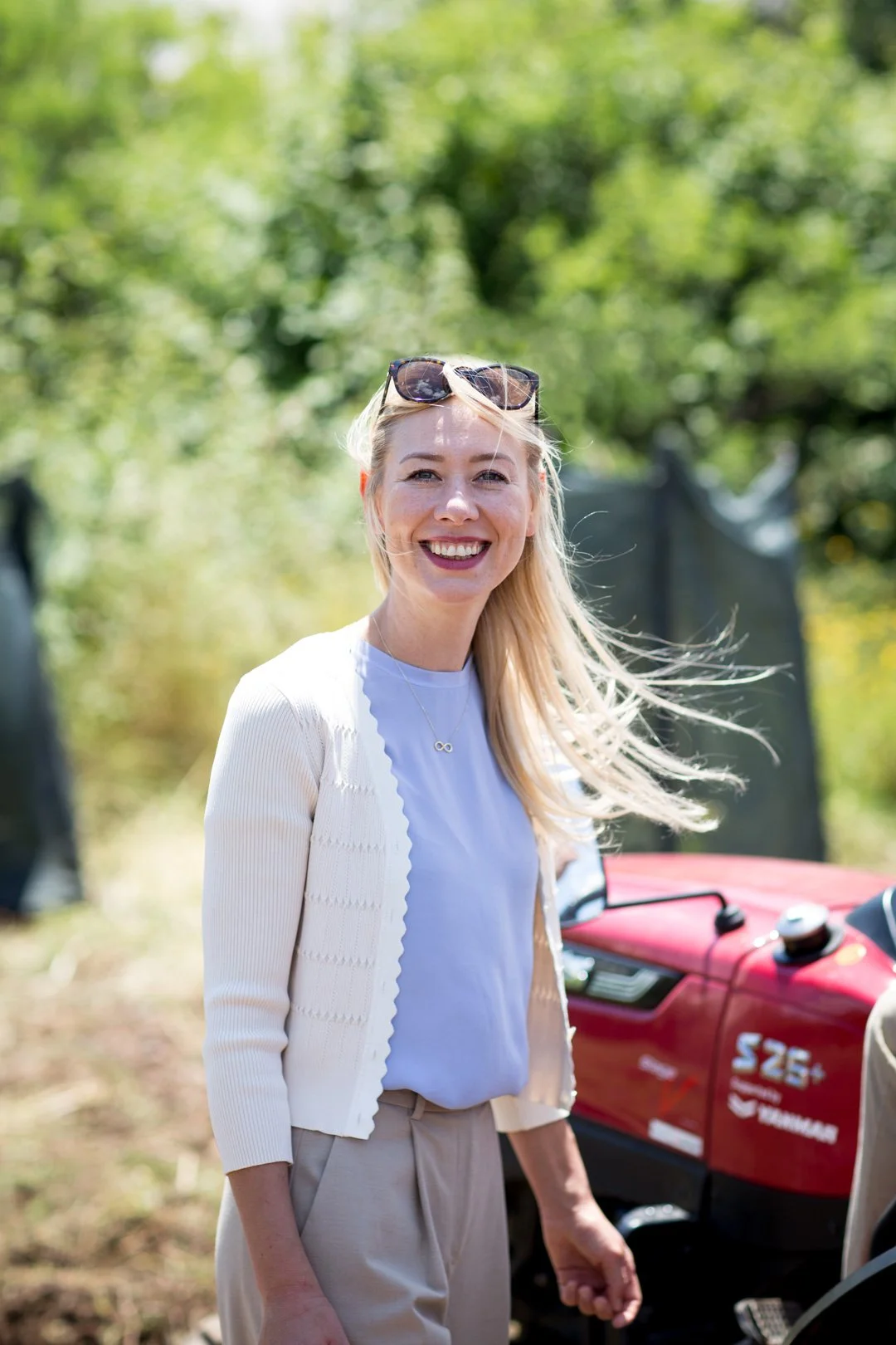 Eine lachende blonde Frau trägt Sonnenbrille auf dem Kopf, einen weißen Pullover und beige Hosen, im Hintergrund sind grüne Bäume und ein rotes Fahrzeug.